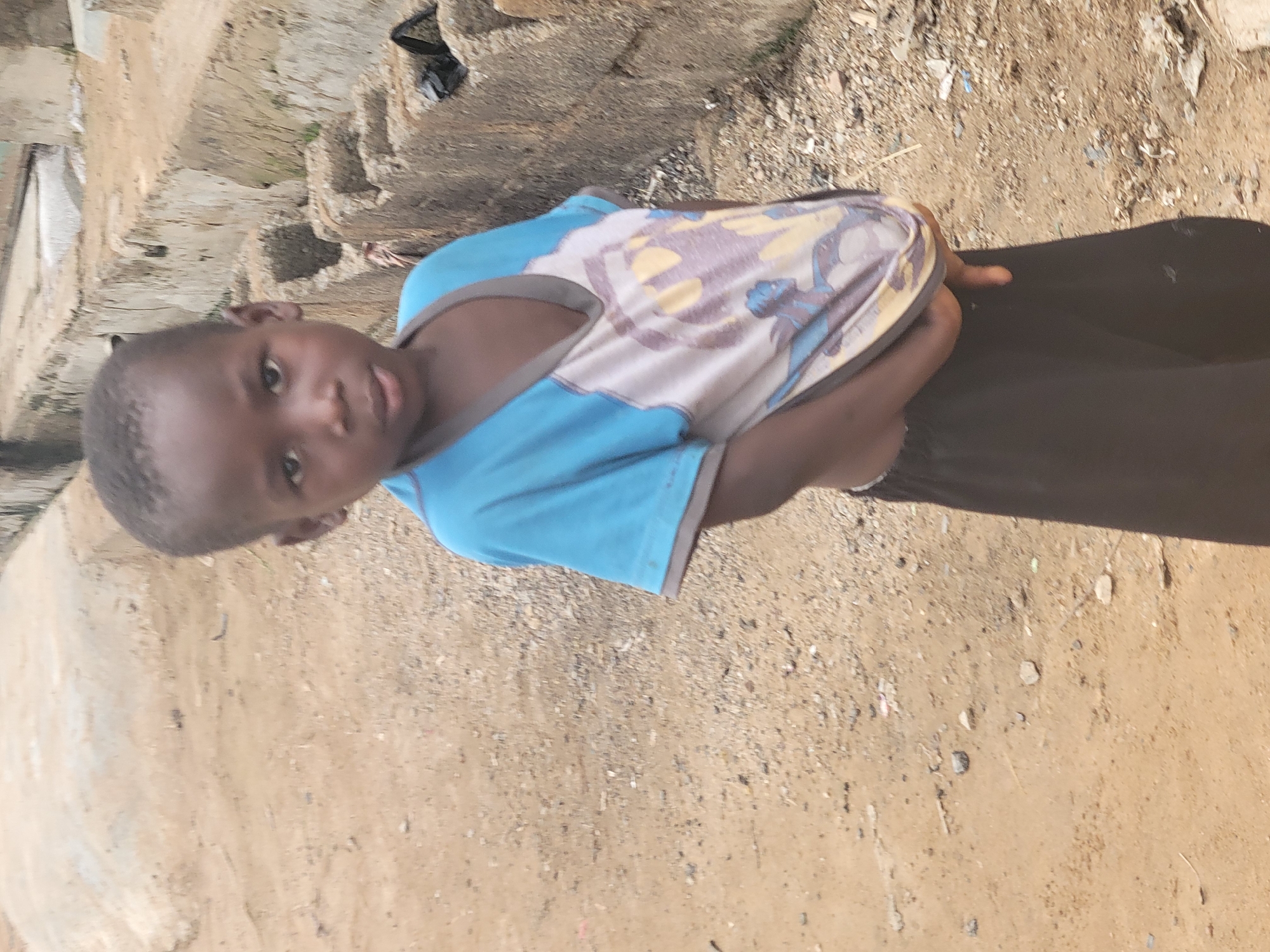 A young child stands on a dusty patch of ground in what appears to be an outdoor location in Madalla, Nigeria. The child wears a short-sleeved shirt with blue and tan/white sections and a dark skirt. The ground is composed of light brown dirt mixed with small rocks and debris. In the background, there is a concrete structure and some trash. The child is looking directly at the camera.