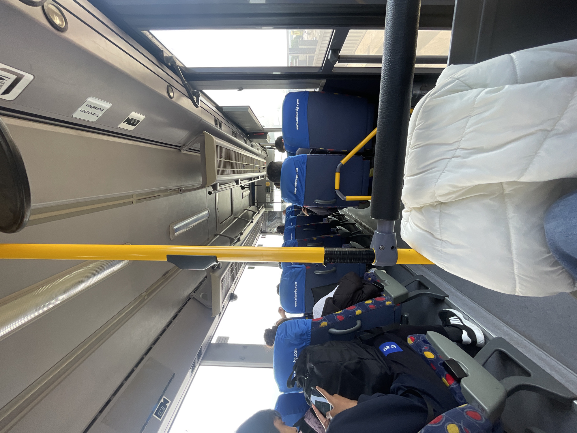 Here is a detailed description of the image:
**Setting:** The interior of a public bus, likely a city bus, captured from an aisle perspective. The image appears to be taken in Unknown, Unknown, possibly in a European country based on the writing and visual cues, but the exact location cannot be determined with certainty.
**Subjects:** The image features numerous passengers sitting along the bus seats. Most individuals are only partially visible, with their heads, shoulders, or backs being the primary visual elements. Details about their age, gender, or specific attire are limited.
**Objects:**
* **Bus Seats:** Most of the seats are covered with blue seat covers, some featuring the website "www.mbus-bg.com".
* **Handrails:** A long, yellow handrail runs along the aisle, providing support for standing passengers.
* **Bags and Personal Items:** Several bags, backpacks, and other personal items are seen on the seats, indicating a mix of passengers possibly commuting or traveling.
* **Clothing:** A prominent white puffy jacket is visible on the right, while other clothing details are obscured by the angle.
* **Bus Interior Elements:** Overhead compartments, interior panels, and window frames are visible, contributing to the overall environment of the bus interior. One of the stickers says "Trittstufen freihalte alten".
**Actions and Interactions:**
* Passengers appear to be sitting passively, potentially commuting or traveling.
* At least one passenger is seen looking at their mobile phone.
* The composition suggests a typical scene of people riding public transportation.
**Environment:** The lighting appears to be bright, possibly due to the windows providing natural light. The environment suggests an urban setting, as buses are a common form of public transport in cities.