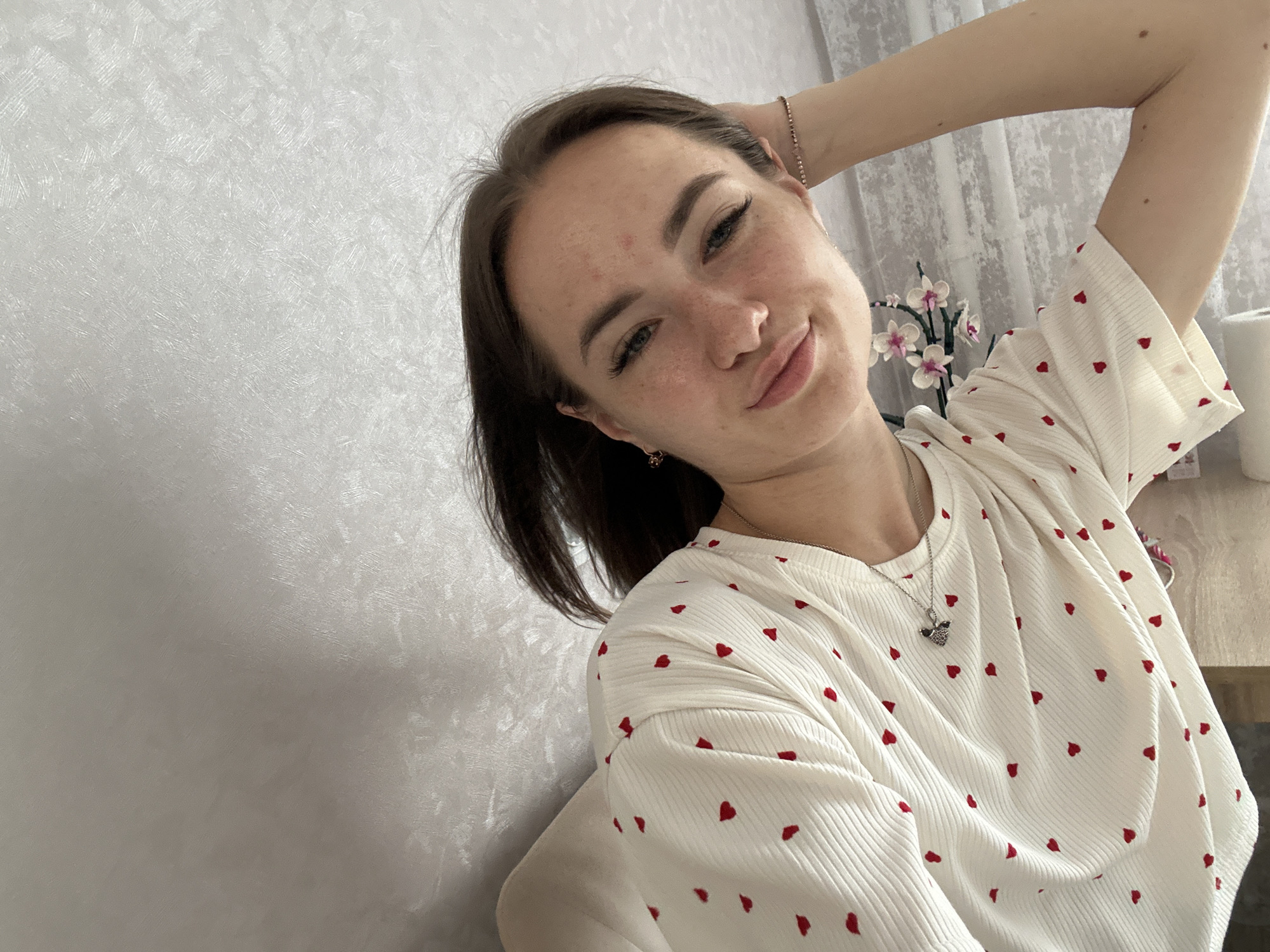 An overhead selfie of a young woman with auburn hair, freckles, and makeup. She is wearing a white ribbed t-shirt with small red hearts and a silver necklace. Her left arm is raised behind her head. She is positioned in the lower right corner of the frame, with a textured white wall filling the left side and a sheer curtain and a plant on a table visible in the background to the right. The setting appears to be indoors, possibly a bedroom, in a location that cannot be determined from the image.