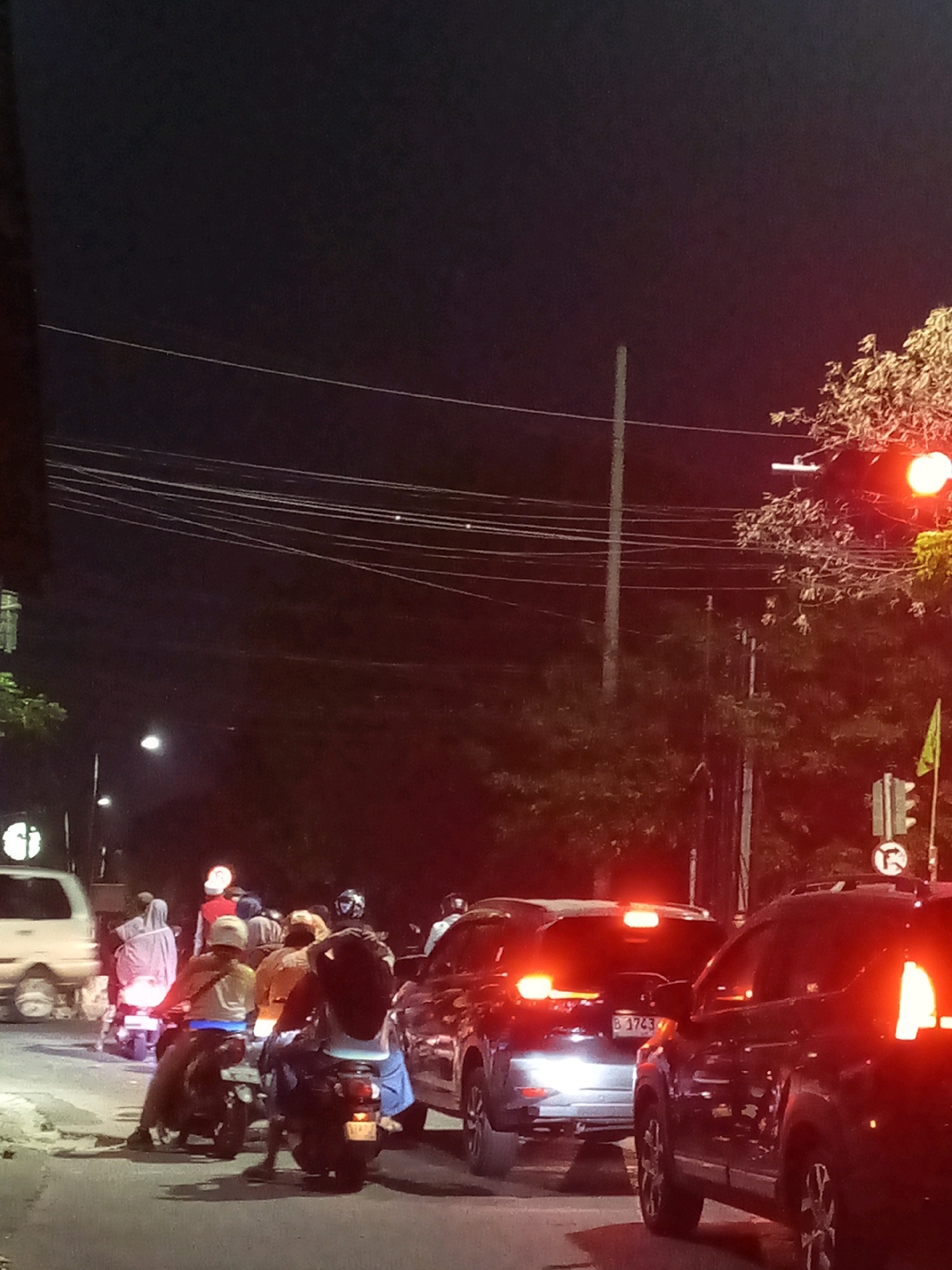 The image depicts a traffic scene at night in Bekasi, Indonesia, likely at an intersection. Multiple vehicles, including cars and motorcycles, are present on a paved road. Several individuals are visible, some riding motorcycles and others possibly as passengers. The vehicles' headlights and taillights are illuminated, contributing to the nighttime ambiance. A traffic light is visible, and it appears to be red. Overhead power lines stretch across the dark sky. Trees and foliage frame the right side of the scene. The overall impression is one of a typical urban traffic scenario at night, characterized by vehicle lights and a flow of movement.
