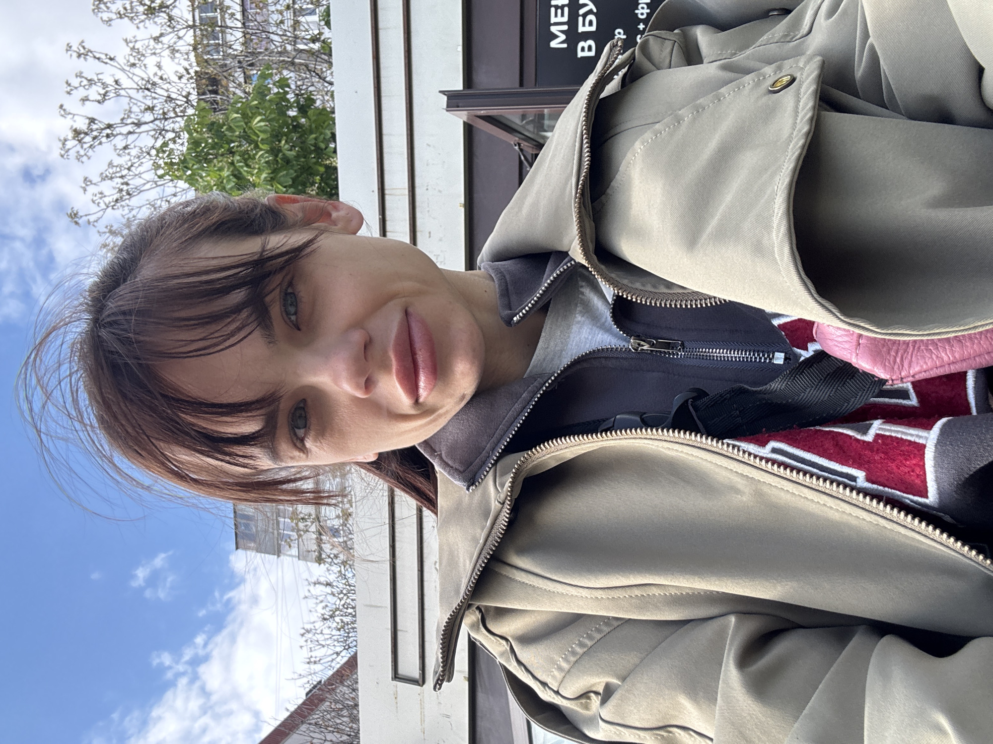 A medium-shot photograph depicts a woman positioned outdoors in what appears to be Rostov-on-Don, Russia. The woman is facing the camera with a slight tilt to her head and a subtle smile. She has brown hair with bangs. She is wearing a light tan jacket with a zipper partially opened to reveal a gray hooded sweatshirt, a garment with red and white details and black straps. Part of a pink object is visible. The background features a partly cloudy blue sky, bare tree branches, and a building with vertical white panels. A sign in Russian with lettering is visible on a dark-colored building.