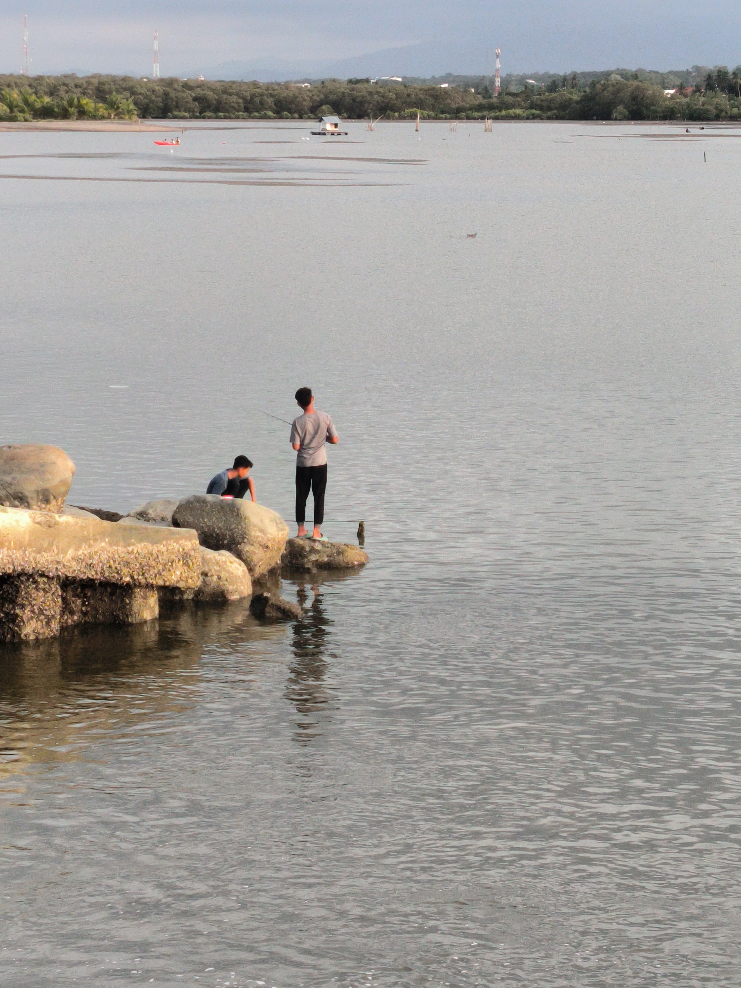 Two young men are on barnacle-covered rocks at the edge of the sea. One is seated and the other is standing and fishing with a pole. In the distance, there are boats and structures in the water. The shoreline in the background is lined with trees, and communication towers are visible beyond the trees. The scene is in Lhokseumawe, Indonesia.