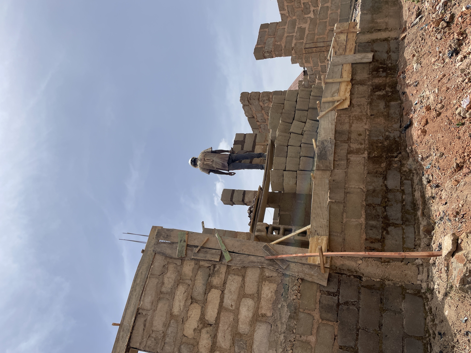 The image depicts an outdoor construction site in Jos, Nigeria, under a light blue sky with scattered white clouds. A solitary male figure, seen from behind, stands on an elevated section of an unfinished building. He wears a light-colored, possibly faded, t-shirt with "IBRAHIMOVIC" and the number "10" printed on the back, dark trousers, and a light-colored head covering. The building is constructed from rectangular gray concrete blocks, with several walls partially erected to varying heights. Crude wooden planks and braces are used as scaffolding and formwork, supporting horizontal structures and a visible stack of additional concrete blocks. Steel reinforcement bars protrude vertically from the top edges of some block walls. The ground beneath the structure is uneven reddish-brown earth, sparsely covered with small rocks and debris. Distant rooftops are visible on the horizon.