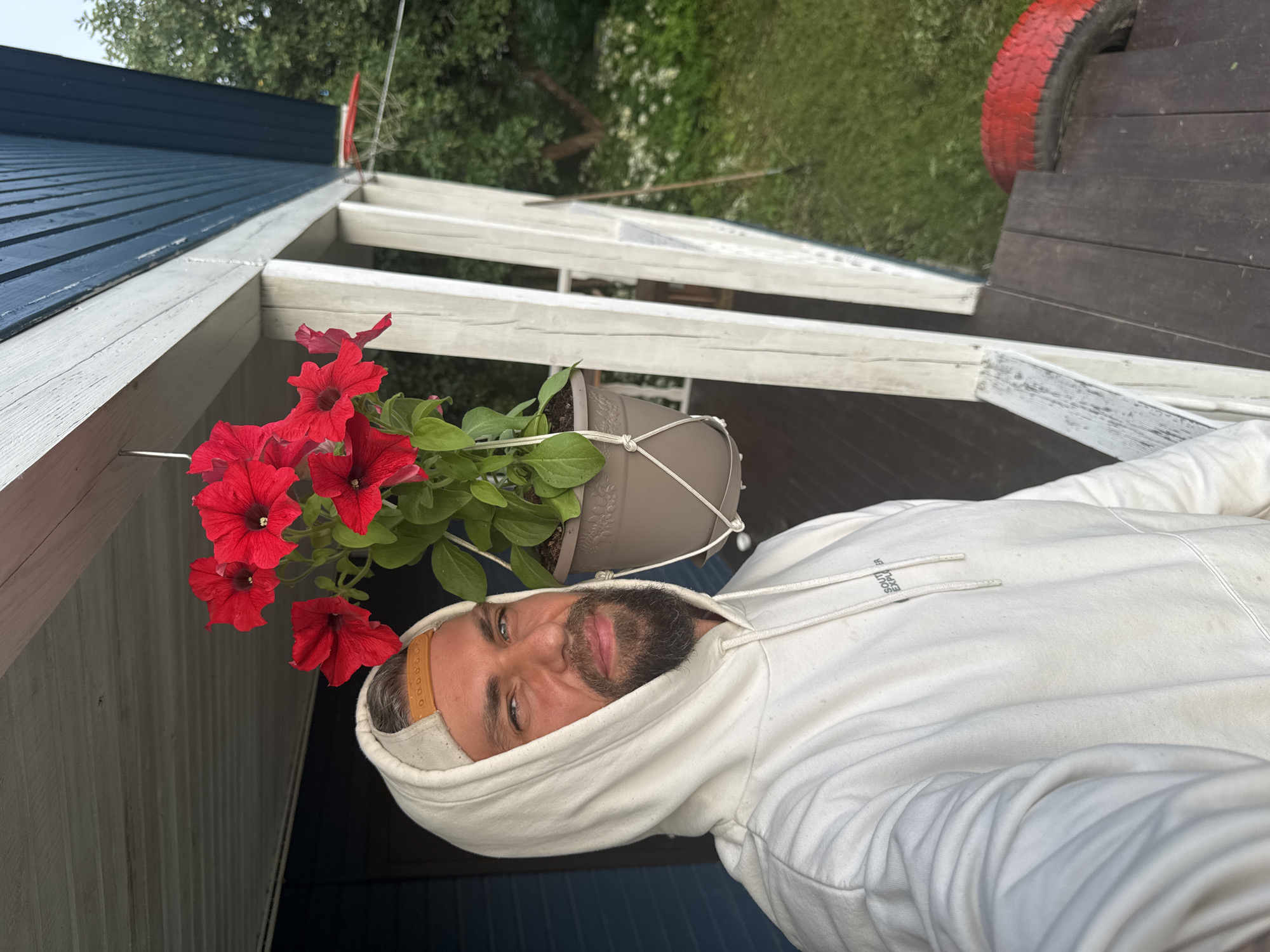 In Kuti, Russia, a man with a beard and wearing a white hoodie and a baseball cap is standing on a porch. He is taking a selfie, and the photo captures his face and upper body. Next to him, hanging from the porch ceiling, is a pot of bright red petunias. The porch has white wooden railings and a dark wooden floor. In the background, a blue wooden house is visible, along with lush green trees and grass. A red-painted tire is also present on the porch.
