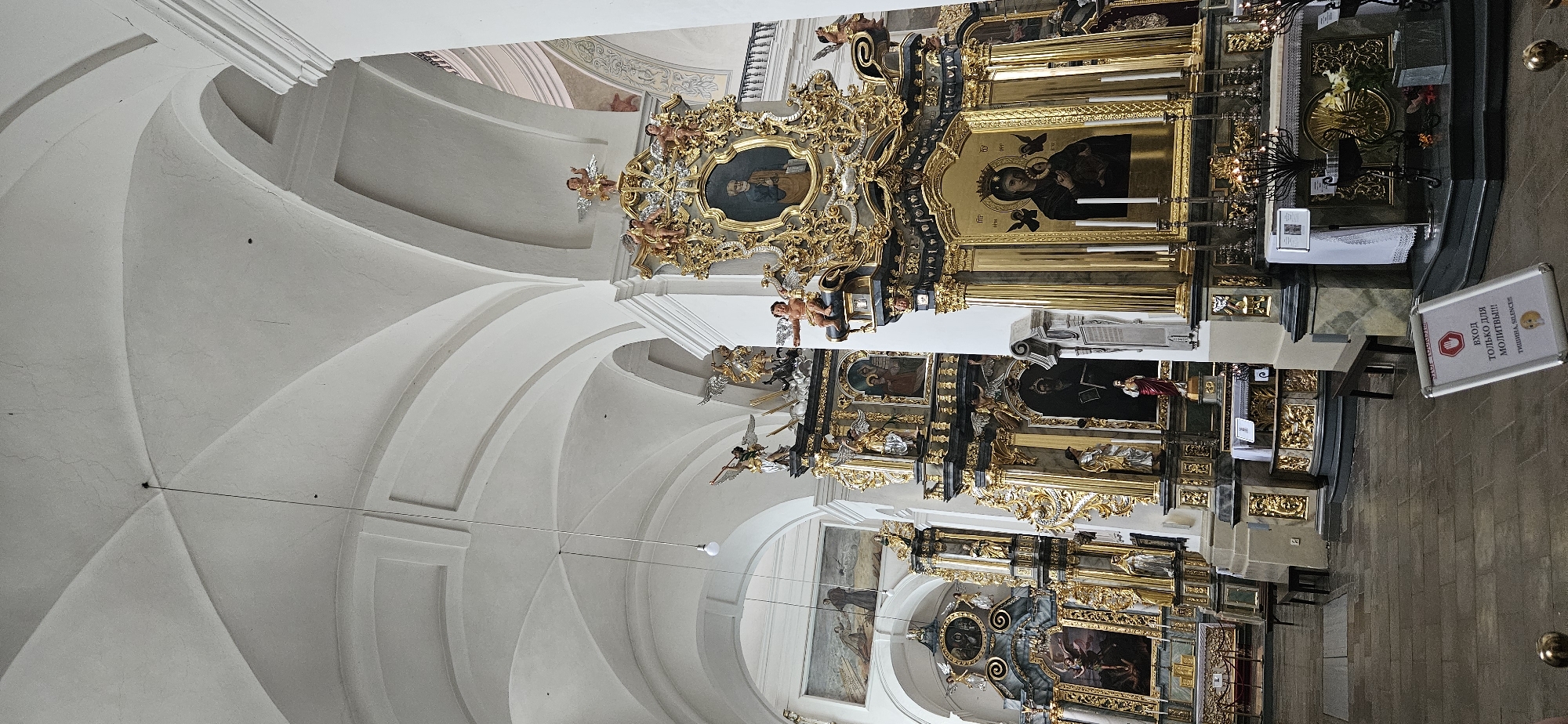 The image shows the interior of a richly decorated church, likely a cathedral, in Hrodna, Belarus. The scene is dominated by ornate gold and black altars, adorned with religious paintings and sculptures of angels and saints. The architecture features high, vaulted ceilings and arched passageways. Several altars are visible, each with multiple tiers of artwork and decorative elements. In the foreground, a sign with Cyrillic text reads "ВХОД ТОЛЬКО ДЛЯ МОЛИТВЫ!!!" which translates to "ENTRANCE ONLY FOR PRAYER!!!" and "ТИШИНА, SILENCE!!!". This sign, along with the overall solemn atmosphere, suggests a place of worship. The floor is made of stone or tile. The lighting appears to be a combination of natural light filtering in and artificial light from lamps and candles. There are no visible people in the immediate foreground or interacting with the altars.