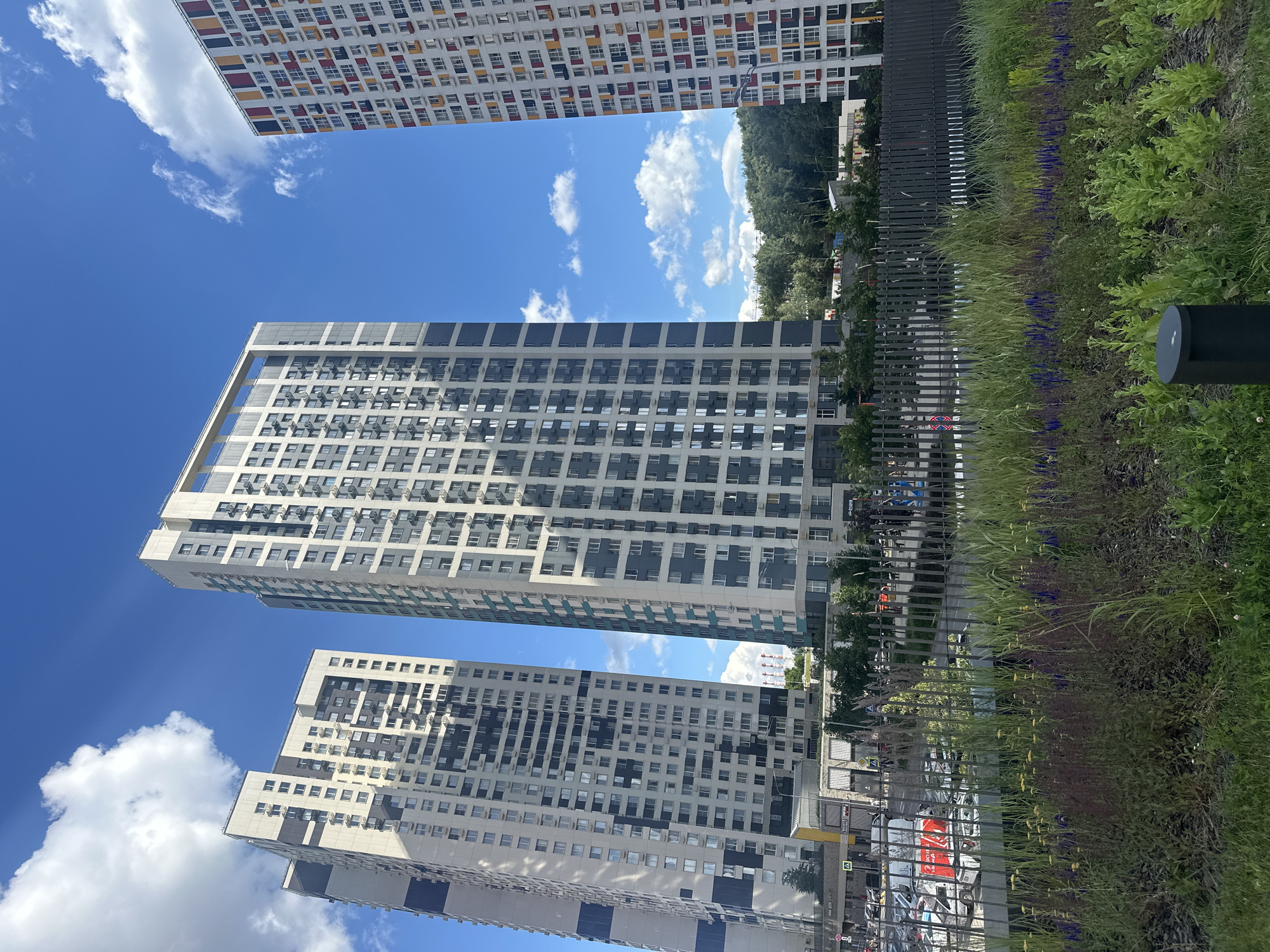 The image shows a bright, sunny day with a clear blue sky dotted with fluffy white clouds. The scene is dominated by several tall, modern apartment buildings. The building in the foreground and middle ground are light-colored with many windows, appearing to be residential blocks. One building in the background has a colorful facade with red, yellow, and white accents on its numerous windows.
To the right of the buildings, there is a lush green area with tall grasses, bushes, and some purple and yellow flowers. A dark metal fence runs along the edge of this green space, separating it from a street or pathway. Some streetlights are visible in the green area, and a dark cylindrical object, possibly another light fixture, is in the lower right corner. In the distance, trees can be seen, suggesting a semi-urban or suburban environment.
The perspective is looking upwards towards the sky and buildings, creating a sense of scale and grandeur. The bright sunlight indicates it is daytime, likely late morning or afternoon, and the clear sky suggests fair weather. There is no visible text in the image. The overall impression is one of a modern, well-maintained residential complex in a pleasant, sunny environment. There are no people or specific activities clearly depicted, suggesting the image captures a static moment of the location.