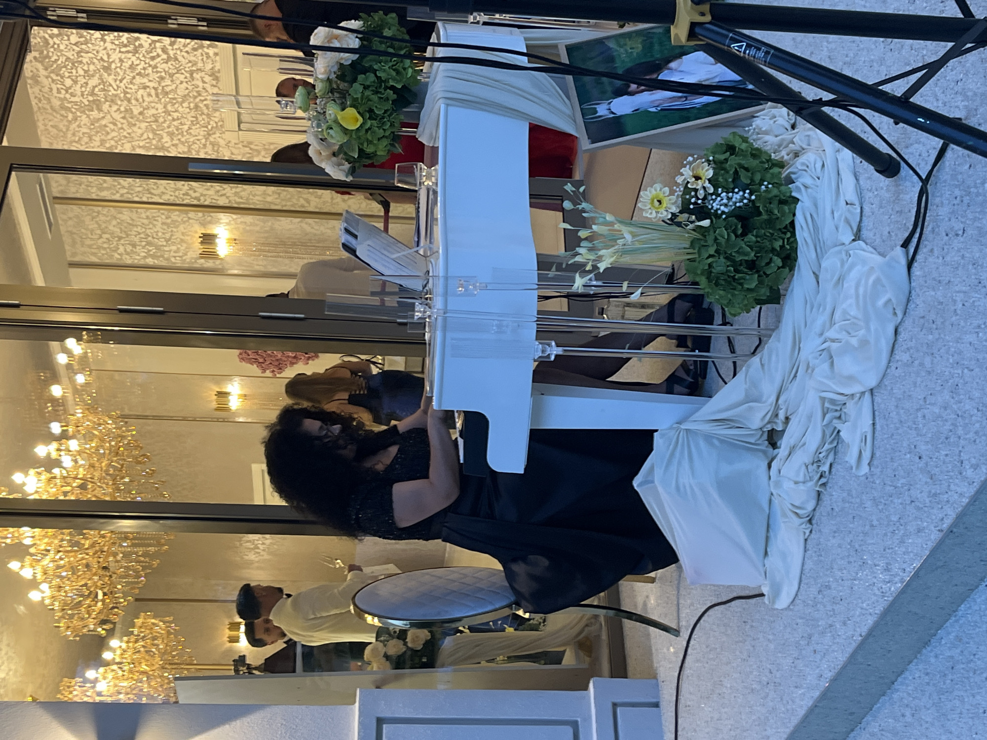 A woman with dark curly hair is playing a white grand piano in front of a large glass-enclosed building. She is wearing a black sequined top and a long black skirt. The piano has a clear stand and sheet music is visible on it. There is a floral arrangement next to the piano, with white and yellow flowers. A framed picture of a white horse is leaning against a speaker stand. The background shows chandeliers and people dressed in formal attire. The ground is tiled and there is a light-colored fabric draped over part of the piano and the floor. The scene appears to be an outdoor event or a building with large open doors.