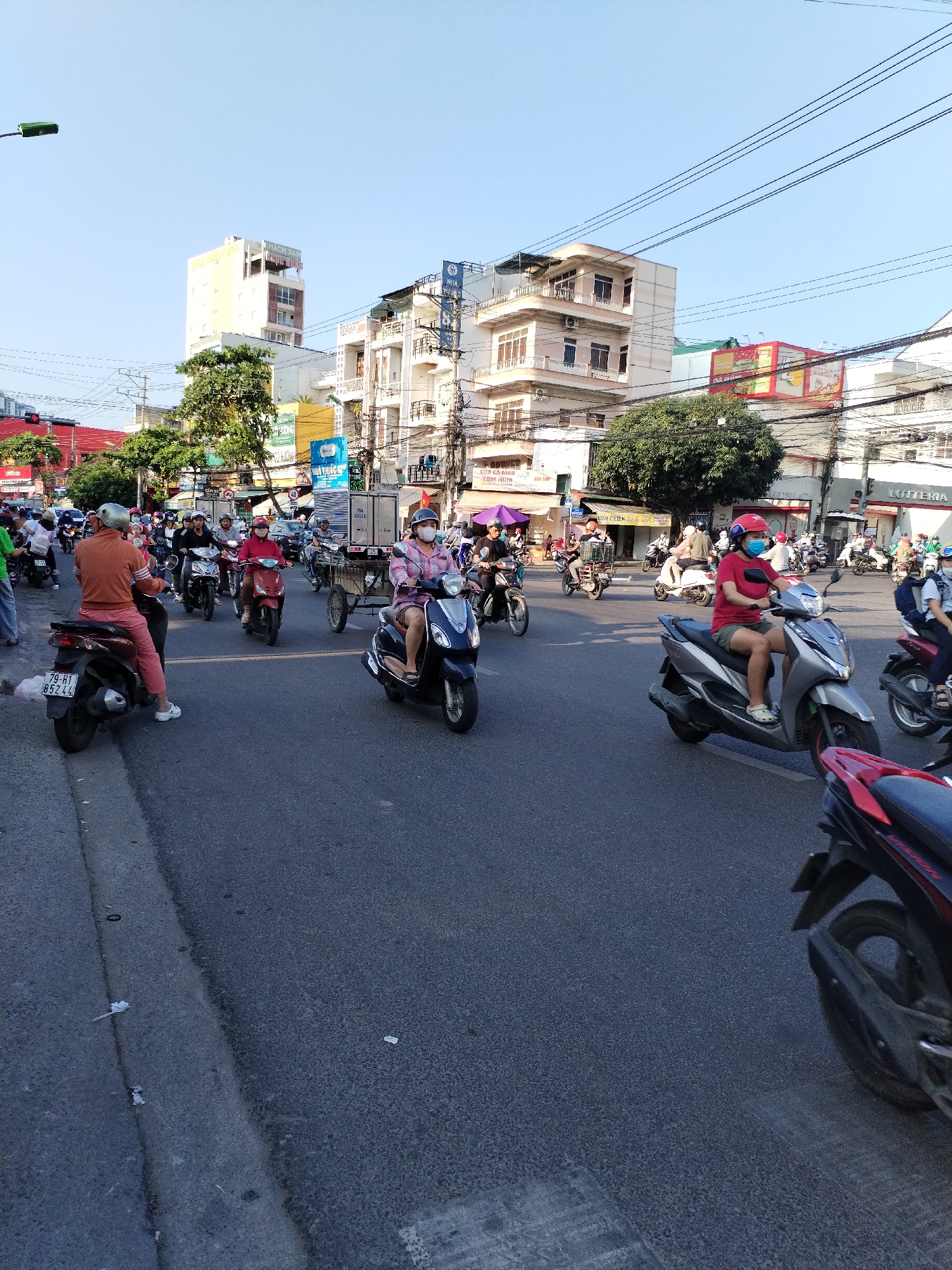 The image depicts a busy street scene in Bac Nha Trang Ward, Vietnam, filled with numerous people riding scooters and motorcycles. The street is paved and the light indicates a bright day. Many riders are wearing face masks. Buildings line the street with various signs and advertisements visible. Utility poles and wires crisscross the sky. In the foreground, parts of scooters are visible, partially obscuring the view. One license plate reads "79-H1 852.44". Other signs are visible, including "LOTTERIA".