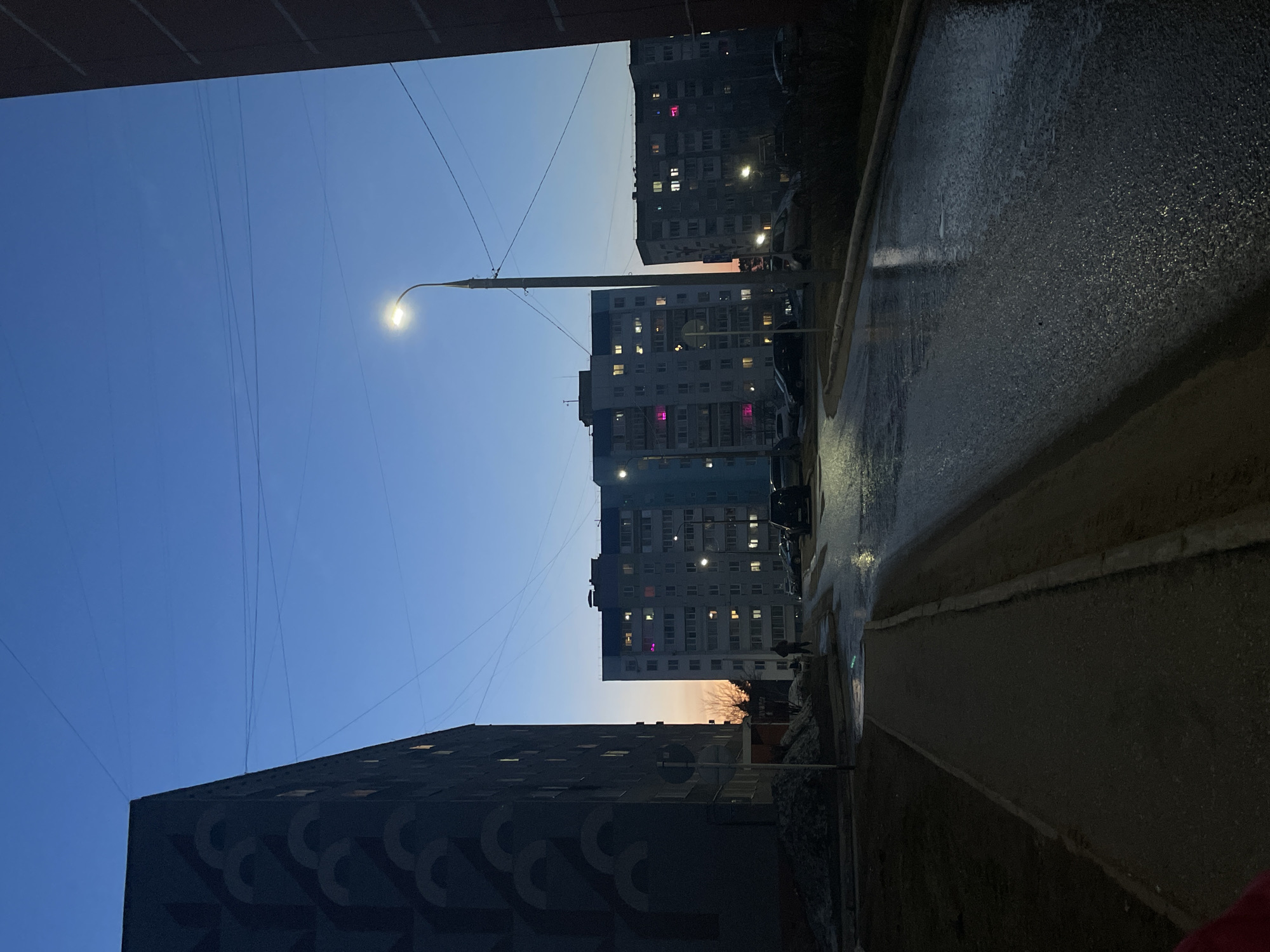 The image depicts a street scene in the city of Chaikovsky, Russia, at dusk or twilight. The perspective is from street level, looking up and between buildings. The buildings are multi-story apartment complexes, common in Russian urban areas. Some windows are illuminated, suggesting occupancy. Power lines are visible against the sky. A street light is on. The ground is wet, likely from recent rain. The road surface reflects the ambient light, creating a sheen. The sky is a gradient of blue to orange, indicating the fading daylight. The image contains no visible people or actions. The environment is urban, suggesting a residential area.