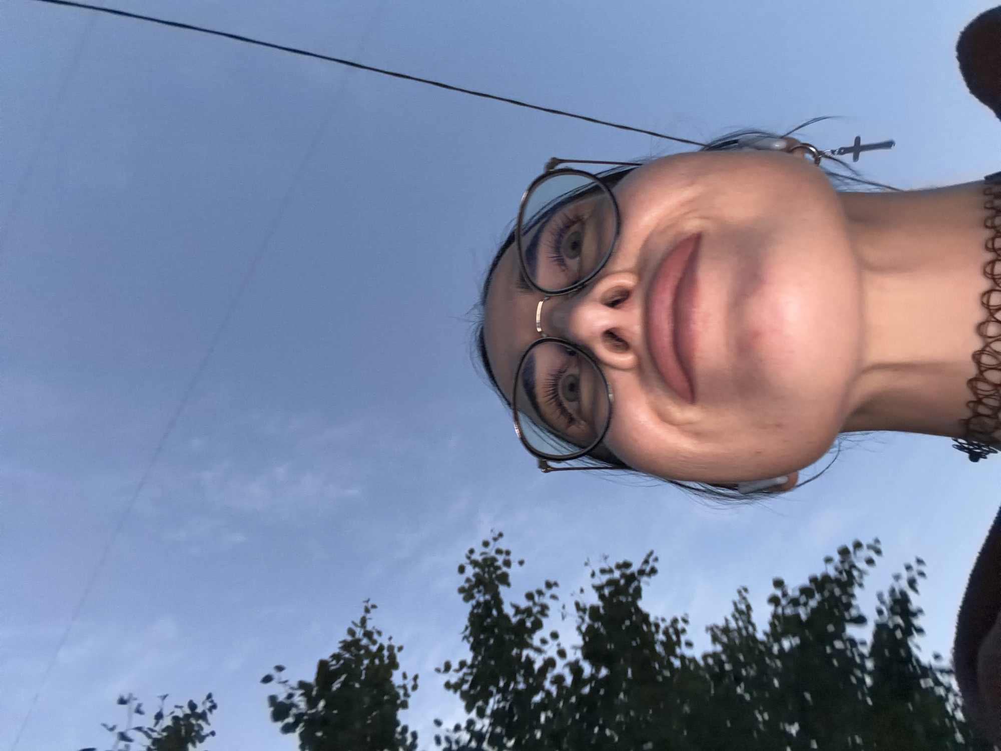 The image is a low-angle portrait, rotated approximately 90 degrees clockwise, capturing a young individual from below looking down towards the camera. The subject, whose head occupies the right side of the frame, wears round, wire-rimmed glasses, a subtle nose ring, and a black, patterned choker. Their eyes are open, revealing light irises, and they have prominent eyelashes. A slight, closed-mouth smile is visible, conveying a pleasant or calm expression. An earring with a small cross charm hangs near their right ear.
The background features a vast expanse of pale blue sky, indicative of dusk or dawn due to its soft, diffused lighting. A thin, dark wire or line diagonally traverses the upper part of the sky. In the lower left quadrant of the image, blurred dark green foliage from trees is visible, suggesting an outdoor setting such as a park or street. There is no discernible text. The perspective creates a candid, upward-looking view, possibly captured in Saint Petersburg, Russia.