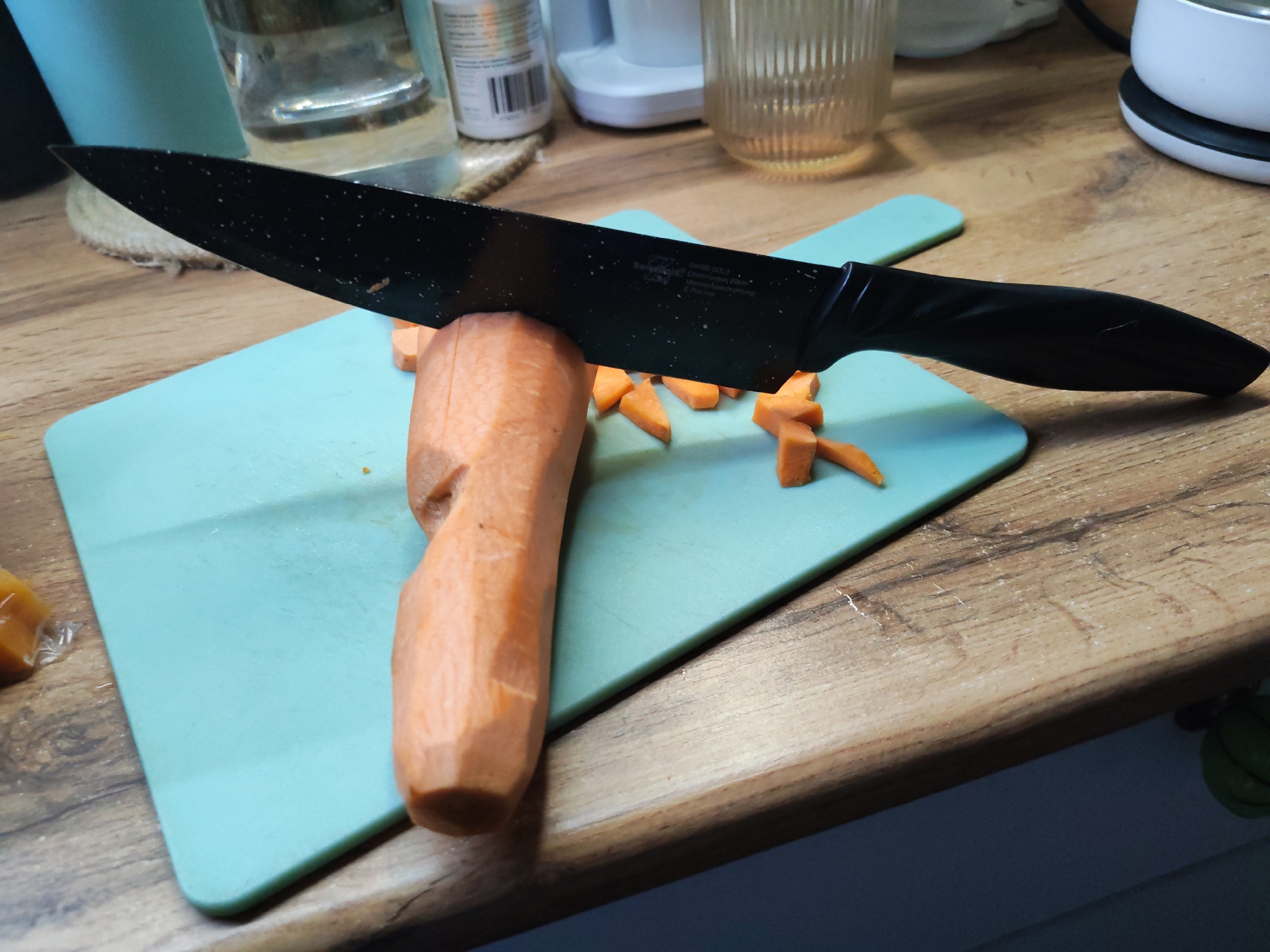 A black knife is cutting a peeled carrot on a light blue cutting board. Several carrot pieces lie on the cutting board to the right of the knife. The cutting board is placed on a wooden countertop. In the background are various items, including a glass container, a white appliance, and a container with a label. The image does not provide information about Izhevsk, Russia.
