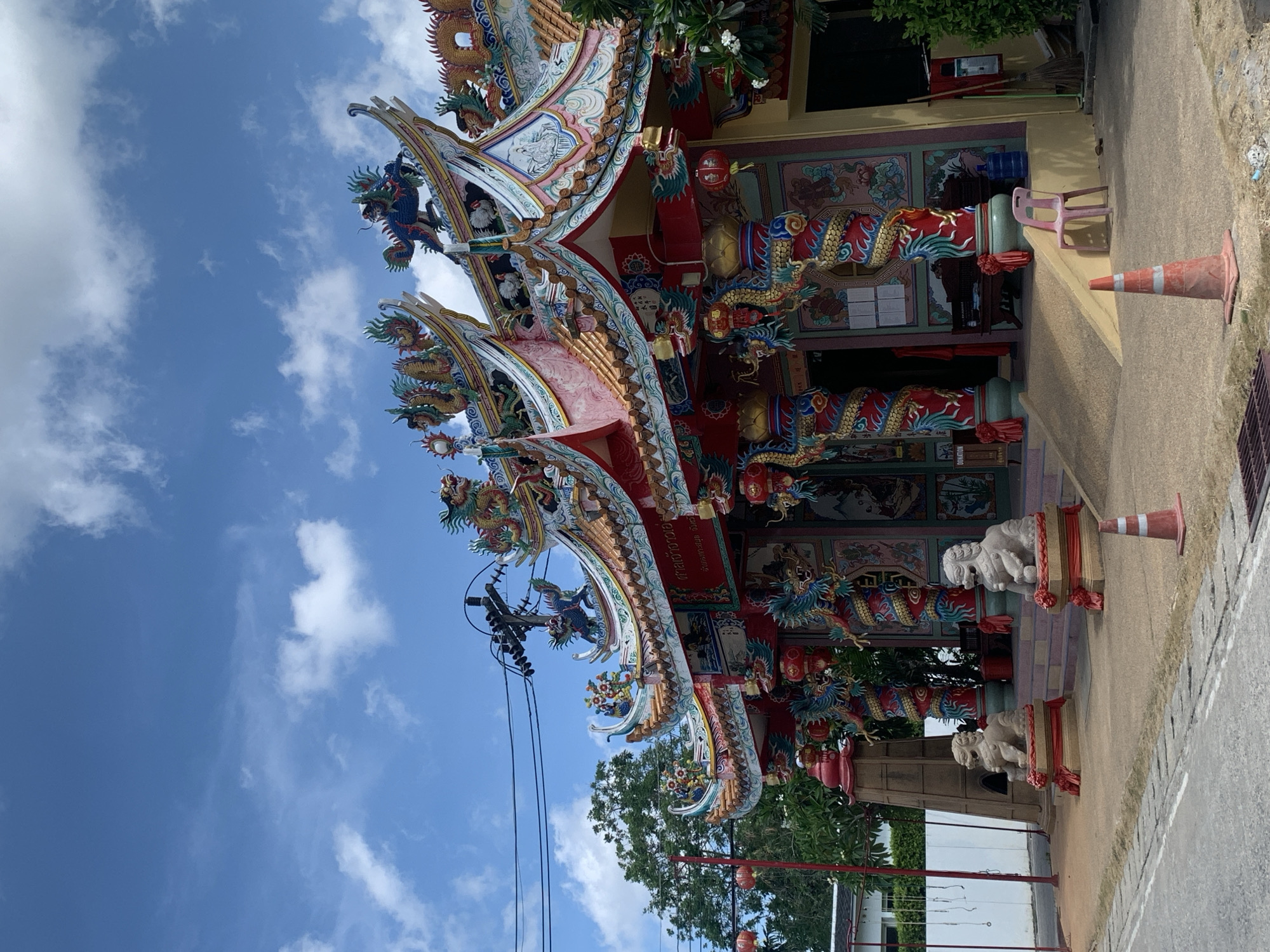 This image captures a vibrant and ornate Chinese temple or shrine, likely located in Baan Mae Nam, Thailand. The photograph is taken from a tilted perspective, looking upwards towards the temple's elaborate roofline and facade against a clear blue sky dotted with scattered white clouds.
The temple's architecture is highly decorative, featuring multiple tiers of gracefully curved eaves adorned with intricate sculptures of dragons and other mythical creatures, rendered in striking colors such as gold, red, blue, and green. Large red pillars wrapped with coiled golden dragons support the structure. The building's entrance displays richly painted murals or detailed relief carvings in a similar vibrant palette, and traditional red lanterns hang from the eaves.
At the base of the temple, two stone lion-like guardian statues (foo dogs) stand on pedestals, each embellished with red sashes. The ground in front of the temple is a paved surface, where a light pink plastic chair and two orange traffic cones are casually placed. Green trees and foliage are visible around the structure, and utility poles with power lines can be seen against the sky on the left.
The scene depicts a bright, sunny daytime, likely morning or early afternoon, with no specific activity or people present. The focus is entirely on the detailed and colorful temple. Visible on one of the temple's architectural elements is text written in Thai script. The image conveys a sense of cultural richness and architectural beauty.