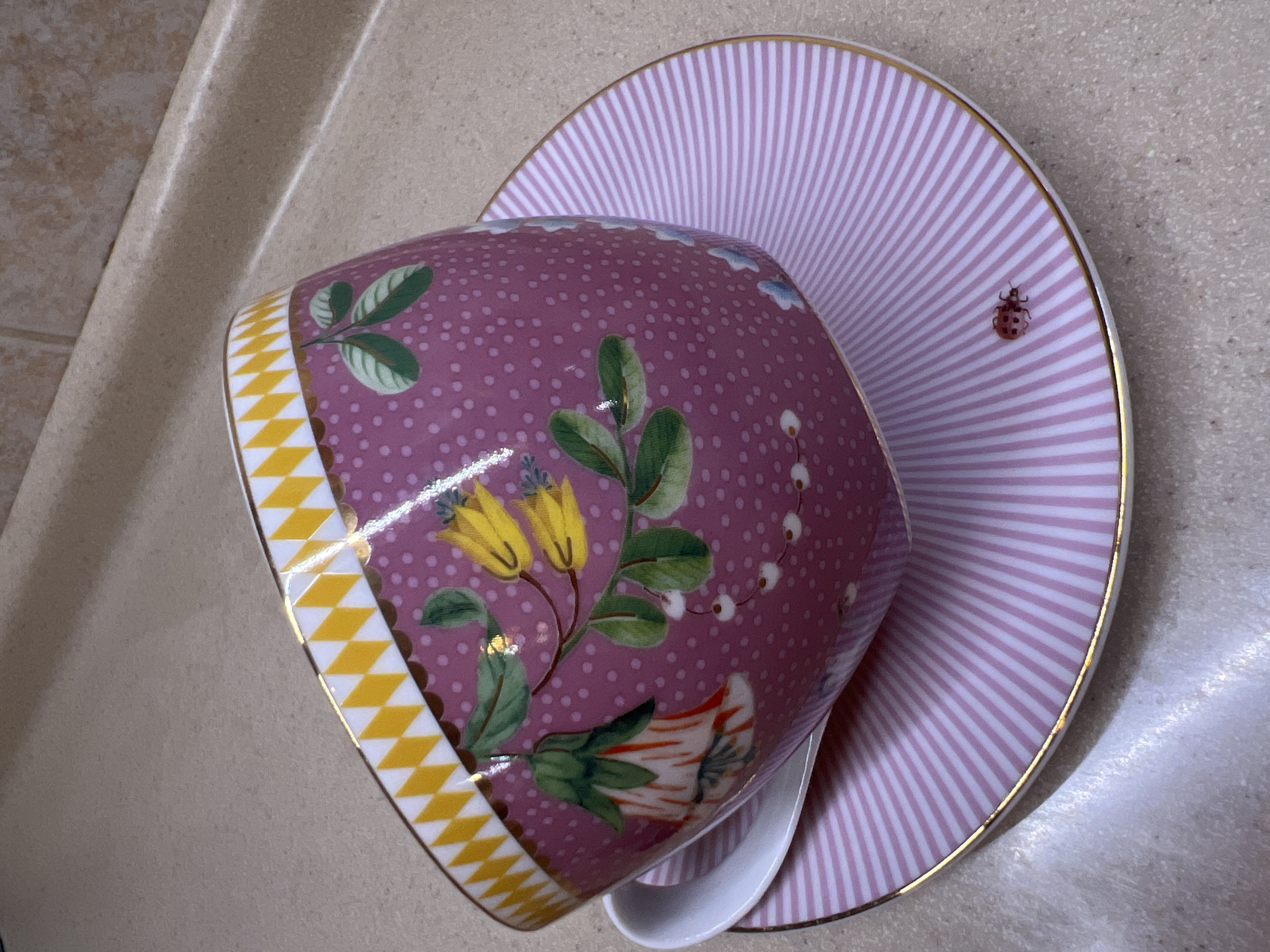 A close-up, high-angle shot captures a decorative tea cup and saucer set on a speckled countertop. The teacup, predominantly pink with small white dots, is adorned with a floral pattern featuring yellow and white bell-shaped flowers, green leaves, and small white berries. A yellow and white diamond pattern borders the rim of the cup. The saucer, also pink, features white vertical stripes and a small, red ladybug design near the outer edge. A white spoon rests partially beneath the teacup. The scene is set in an indoor environment, possibly a kitchen or dining area, with a subtle hint of a tiled backsplash in the background. The location is not Saint Petersburg, Russia.