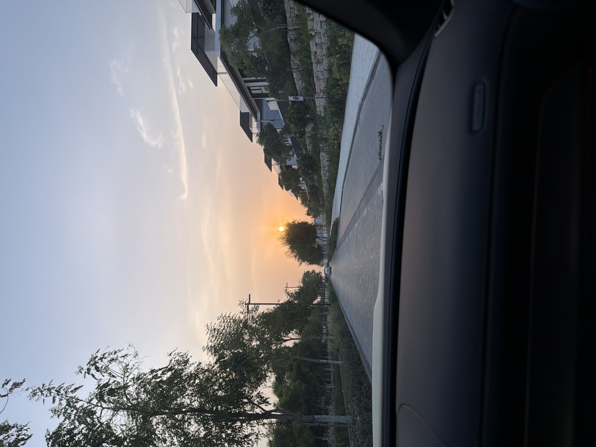 The image captures a view from inside a car, looking out onto a paved road in Dubai, United Arab Emirates, during a sunset. The road, lined with trees and modern residential buildings, is illuminated by the warm, orange glow of the setting sun. The sky transitions from a clear, pale blue at the top to a soft orange and yellow hue closer to the horizon. The buildings, characterized by their sleek, contemporary architecture with flat roofs and large windows, are situated on an elevated, terraced landscape. The foreground is dominated by the car's dashboard and windshield, suggesting the perspective of a driver or passenger. The overall scene conveys a sense of tranquility and urban sophistication.