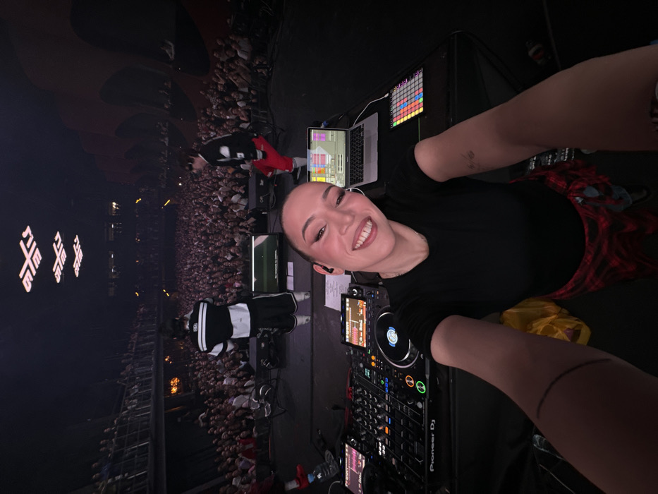 A wide-angle selfie depicts a live concert scene at Adrenaline Stadium in Moscow, Russia. In the immediate foreground, a young adult female, identified as the photographer, smiles directly into the camera while positioned behind a professional DJ setup. She wears a black t-shirt and has a red and black plaid shirt tied around her waist. Her right arm is extended, holding the camera. The DJ console includes a Pioneer DJ mixer, two Pioneer CDJ players, an open silver MacBook displaying music production software, and a multicolored grid controller.
On the main stage, beyond the DJ setup, two male performers are visible, facing a large, densely packed audience. One performer wears a black and white athletic top and black shorts with white stripes. The other wears a black and white top with bright red pants. Two large dark screens are positioned on the stage floor behind them.
The audience, a vast crowd, fills the main floor and a multi-level balcony section on the left side of the venue. Many individuals are holding up mobile phones. The venue's ceiling features three prominent, illuminated white stylized 'X' symbols, characteristic of Adrenaline Stadium. The side walls exhibit a distinct ribbed architectural design, with warm orange lighting accents visible on the right. The overall lighting is dim, emphasizing the stage and ceiling symbols.