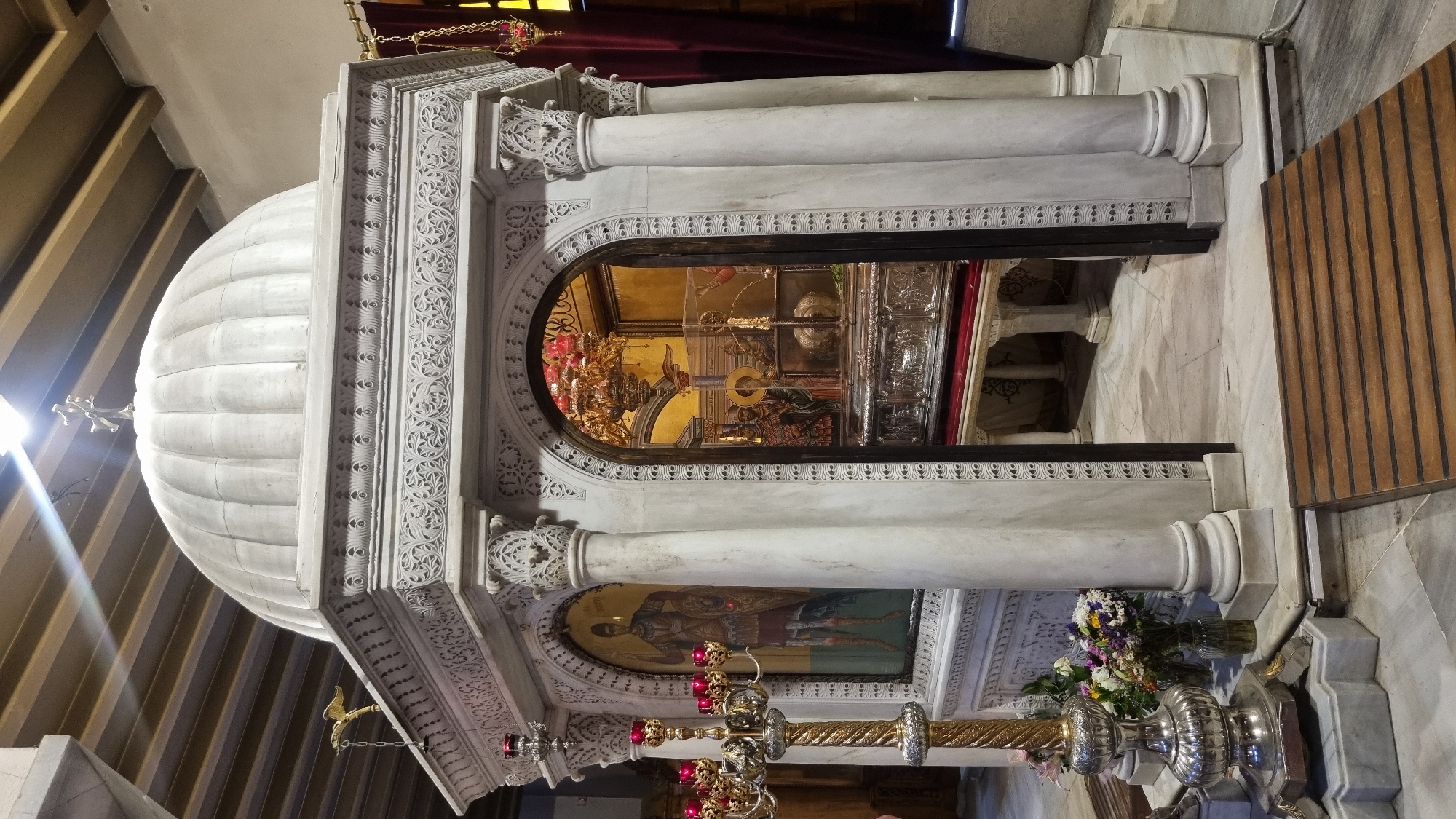This is a full shot of a marble structure in Thessaloniki, Greece, containing religious artifacts. The structure is ornate, with a dome on top topped with a cross. Carvings adorn the sides of the structure, and pillars support the roof. Inside the structure, visible through an arched opening, is a silver reliquary adorned with intricate carvings, resting on a red velvet cloth atop a wooden table. Several religious icons are also visible inside, including one depicting a warrior saint. A large, ornate candelabra with red candles stands to the left of the structure, with a vase of flowers on the floor nearby. A wooden ramp leads to the entrance of the structure. The ceiling, made of wooden beams, is visible at the top of the frame. The floor is tiled marble. The scene appears to be indoors, likely in a church or a religious shrine.