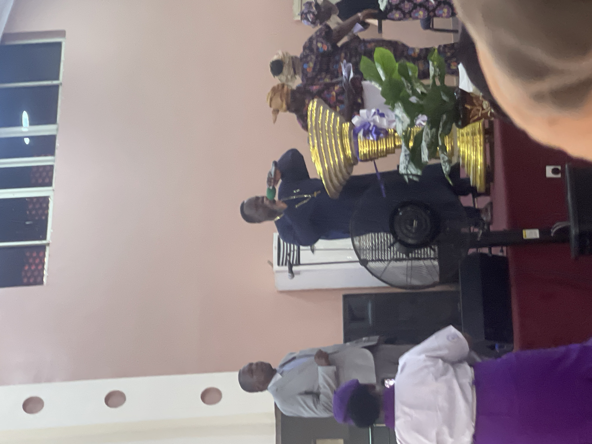 The scene appears to be inside a building in Kubwa, Nigeria. A man in a dark blue suit is holding a microphone and addressing a group of people. Behind him are two women in traditional clothing and a gold-colored decorative object with white flowers. A standing fan is also behind the man. In the foreground, there is a man in a grey suit and two people wearing white and purple clothing. The room is painted in a light pink color, and there is a window with a grid pattern on the left side of the image. A dark-colored speaker is visible in the background.