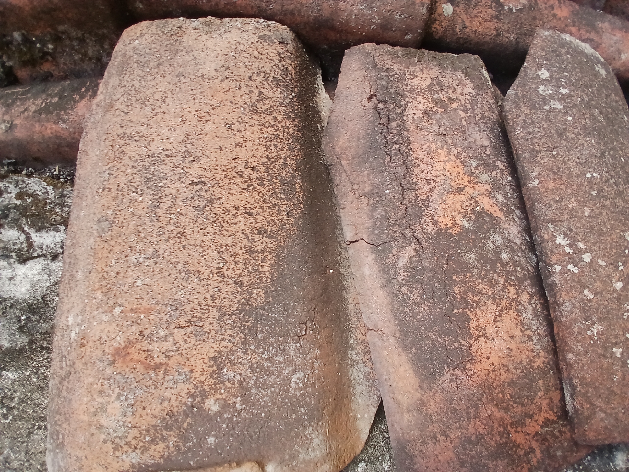 A close-up, overhead view shows a stack of weathered, reddish-brown clay roof tiles. The tiles are rough in texture and show signs of age and exposure to the elements, with patches of darker discoloration and lighter mineral deposits. The arrangement suggests they are either in storage or part of a rooftop structure. The image is devoid of human presence. The location is the city of Kademangan Kulon, Indonesia, but specific details about the precise setting within the city are not provided by the visual information.