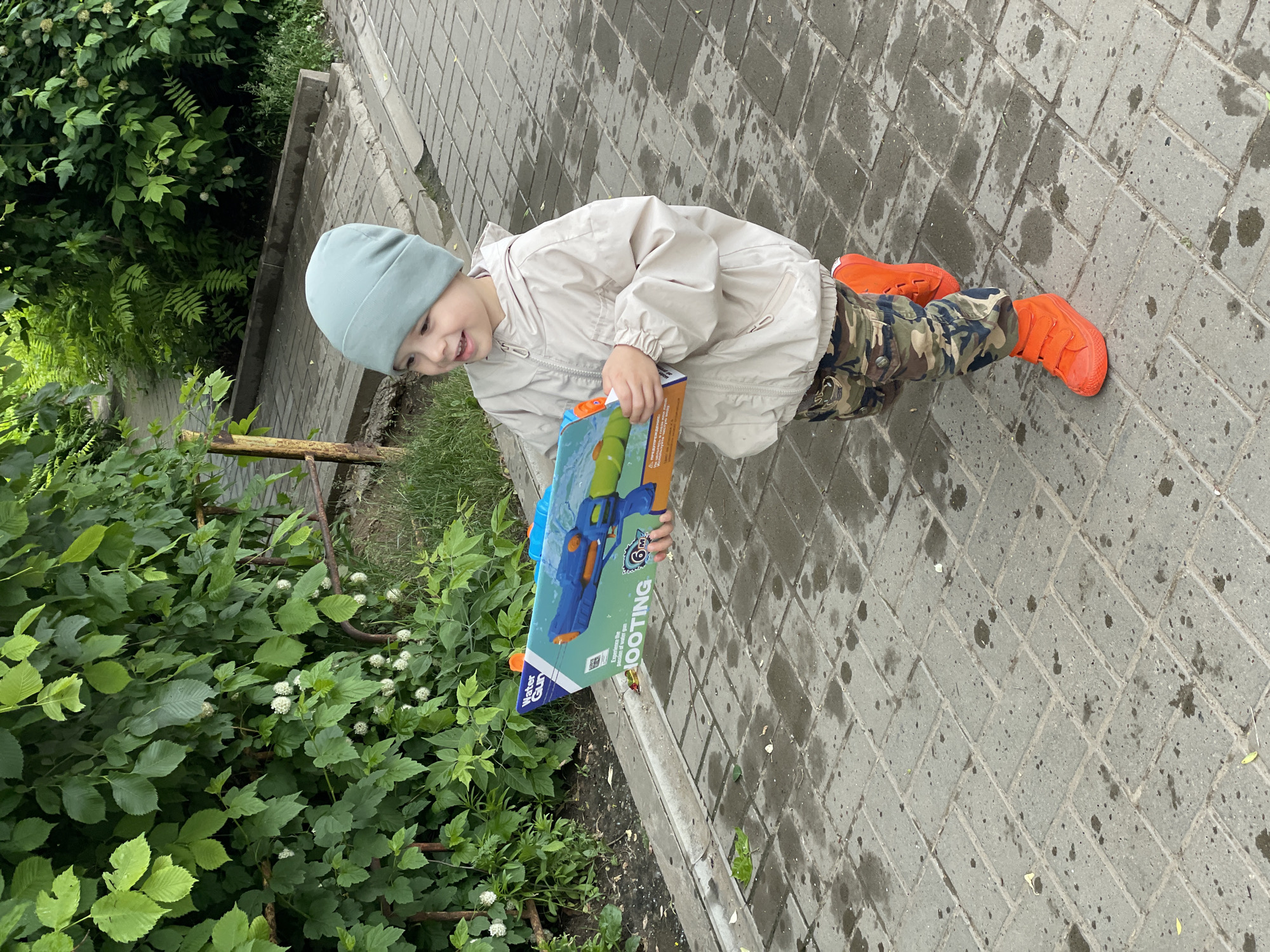 A young child is captured in an outdoor setting, smiling brightly while holding a packaged toy water gun. The child is dressed in a light blue or mint green beanie, a light beige jacket, camouflage-patterned trousers, and striking bright orange shoes. Their expression conveys happiness and excitement, likely over the new toy.
The scene features a grey paved path, composed of rectangular blocks, which appears wet in various spots, possibly from recent rain or water play. To the left of the path, there is a dense cluster of lush green foliage, including bushes with small white, spherical flowers. Some bare earth is visible beneath the plants.
The toy water gun packaging is prominently displayed, featuring a blue and green water gun design. Visible text on the packaging includes "WATER GUN," "SHOOTING," and "6M," suggesting its type and range. The overall impression is of a joyful moment, perhaps after receiving a new toy, with a hint of damp weather or recent water activity. The lighting suggests it is daytime.