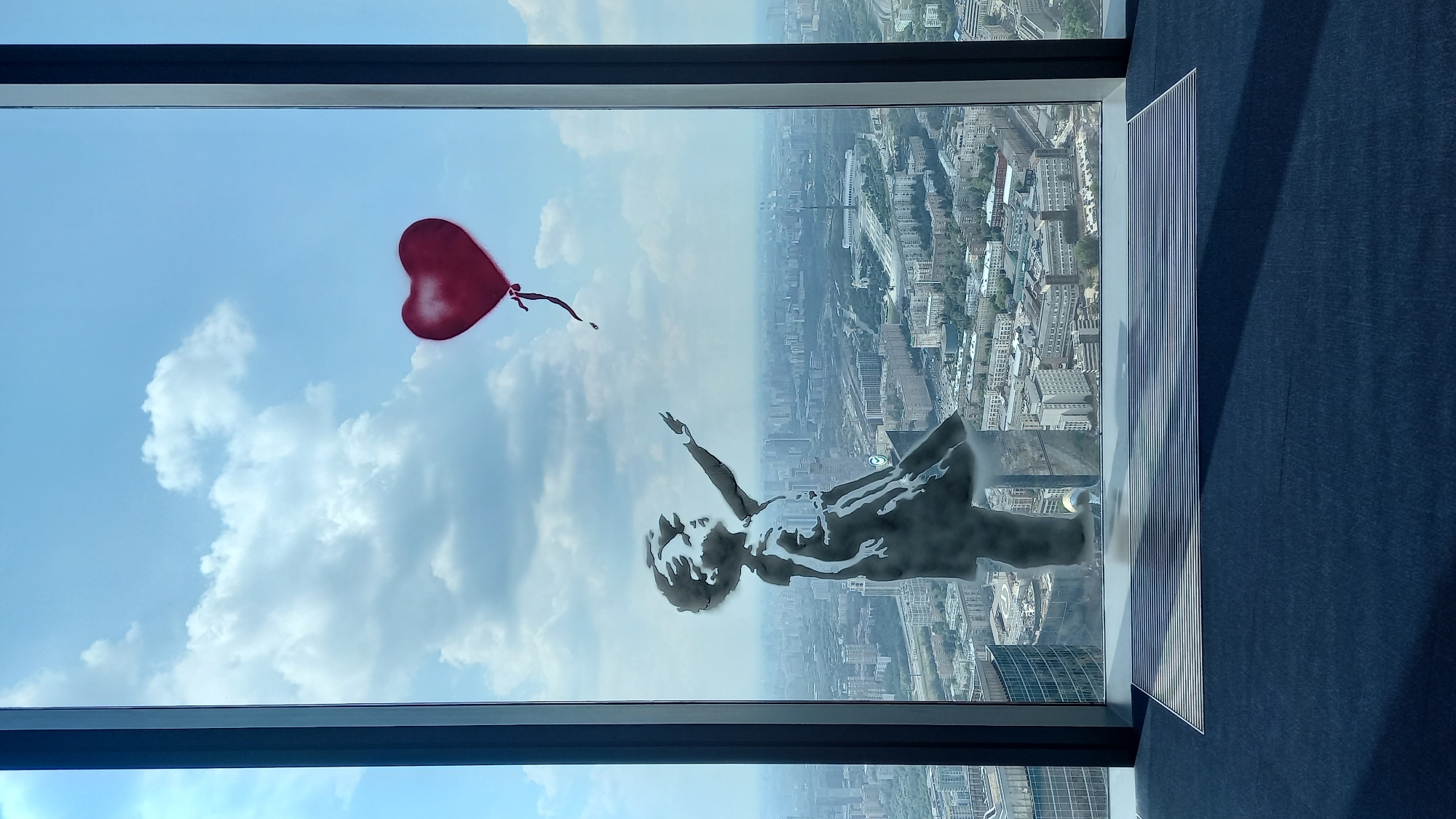 The image shows a stencil of a child reaching for a red heart-shaped balloon, applied to the inside of a large window. The stencil is a silhouette in black and white, with some shading. The child's arm is extended upwards, and their fingers are reaching towards the balloon. The balloon is bright red and appears to be floating away from the child.
The window overlooks a sprawling cityscape under a cloudy blue sky. The city is densely packed with buildings of various sizes and architectural styles. In the foreground, the window frame is dark and prominent. Below the window, a dark blue carpet covers the floor, with a striped metallic mat near the window. The overall impression is one of a poignant artwork displayed in a high-rise setting, juxtaposing a child's hopeful gesture with the vastness of the urban environment. The location is identified as Unknown, Unknown.