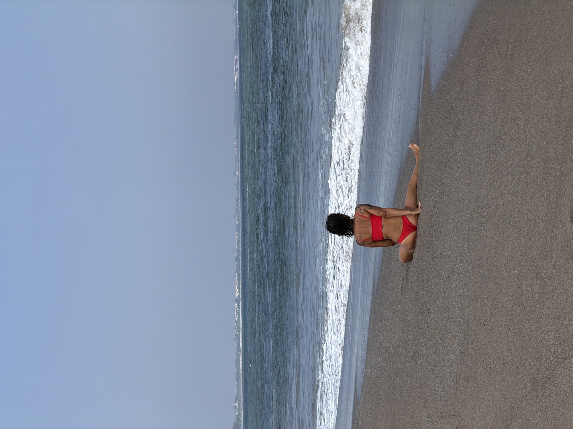 A woman in a red bikini sits on a sandy beach, facing the ocean. She has dark hair and tattoos on her arm. The water is a deep blue with white waves breaking near the shore. In the distance, a hazy coastline with buildings is visible under a pale blue sky. The image appears to have been taken in North Kuta, Indonesia, likely in a popular beach area.