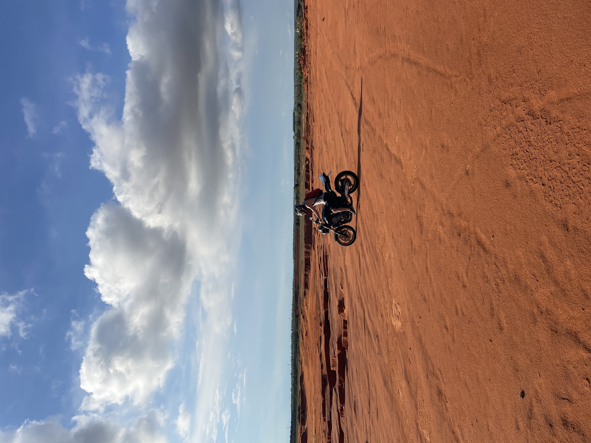 A person is riding a motorcycle across a sandy, orange-colored landscape with stratified rock formations in the background. The rider, wearing a helmet and a white tank top, has a red backpack. The sky above is blue with scattered white and gray clouds. The scene appears to be a desert or badlands environment, consistent with the red sand dunes of Mui Ne, Vietnam. The motorcycle is black and gold and is angled to the left of the frame. The terrain is uneven with tire tracks visible in the sand.