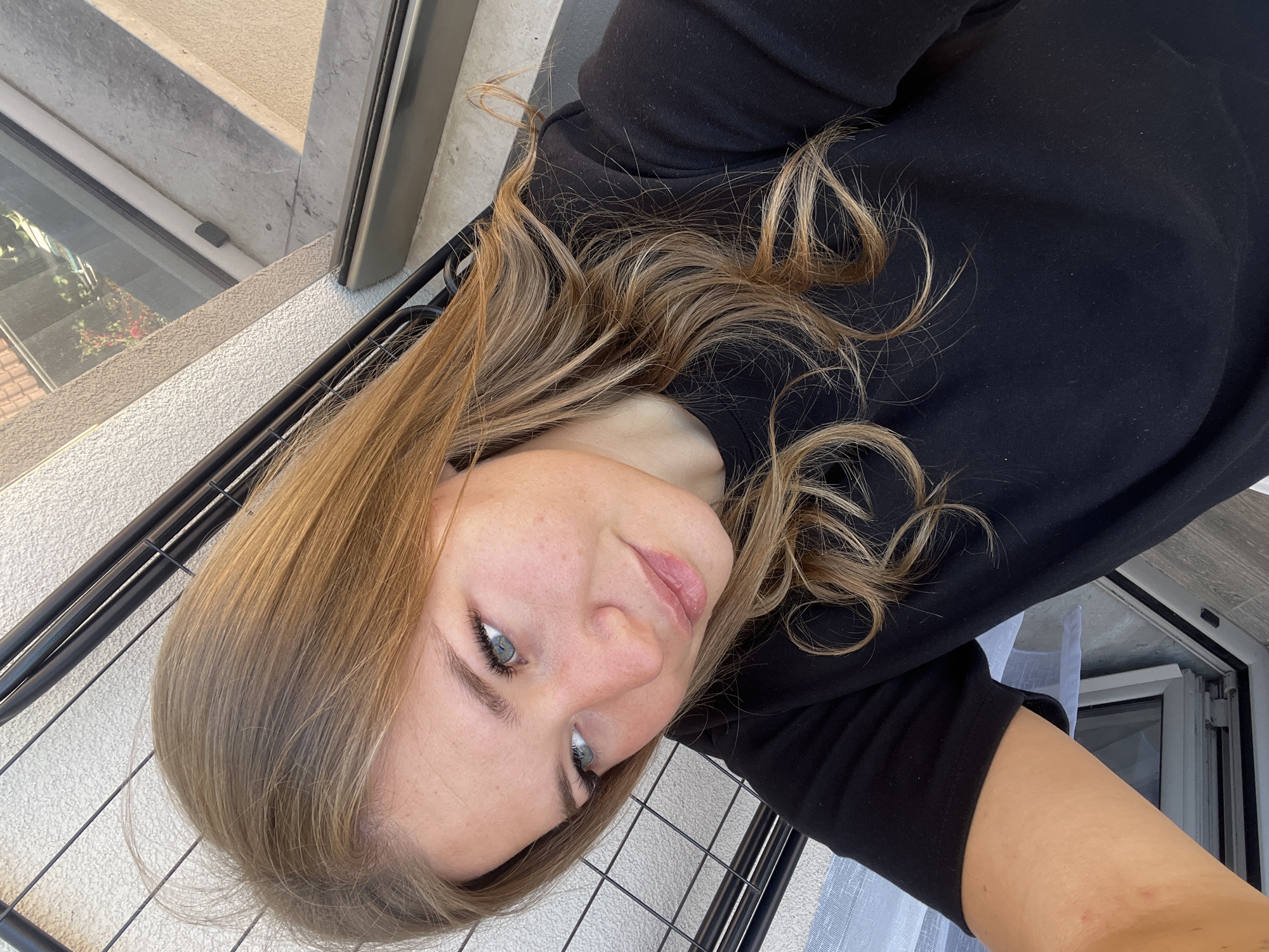 A selfie-style photograph shows a woman with blonde hair, wearing a black hoodie. She is positioned at an angle, with a metal railing visible behind her, suggesting a balcony setting. In the background, there is a partially visible building exterior and window, which could be a feature of Budva, Montenegro. The photo is taken from a slightly elevated perspective.