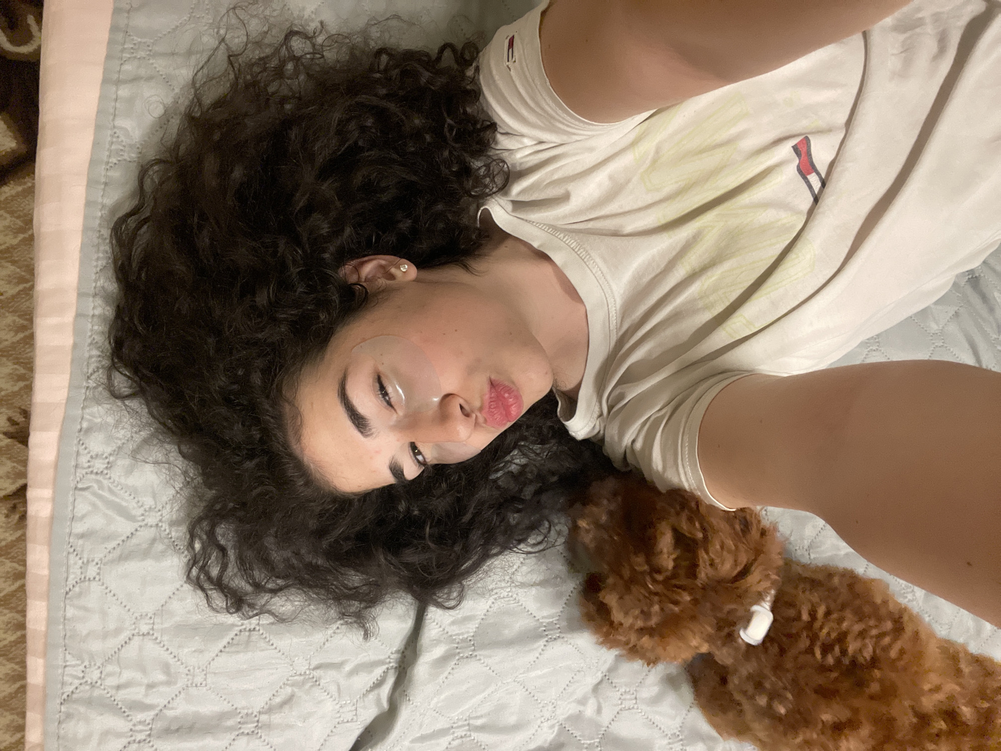 A young woman with dark, curly hair is lying on her back on a light blue, quilted comforter, taking a selfie with her right arm extended. She is wearing a white t-shirt with a graphic design. Under her eyes, she has light-colored eye patches. Her lips are pursed, and her left eye is slightly squinted. Next to her, to her left, a small, reddish-brown toy poodle is lying down, its head resting on the woman's arm. The dog appears to have a white object attached to its collar. The background shows a bed with a textured comforter and a strip of fabric with a pattern of brown, beige, and pink lines at the top of the image. The scene appears to be indoors, in a bedroom, with no specific visual indicators pointing to the city of Sofia, Bulgaria.