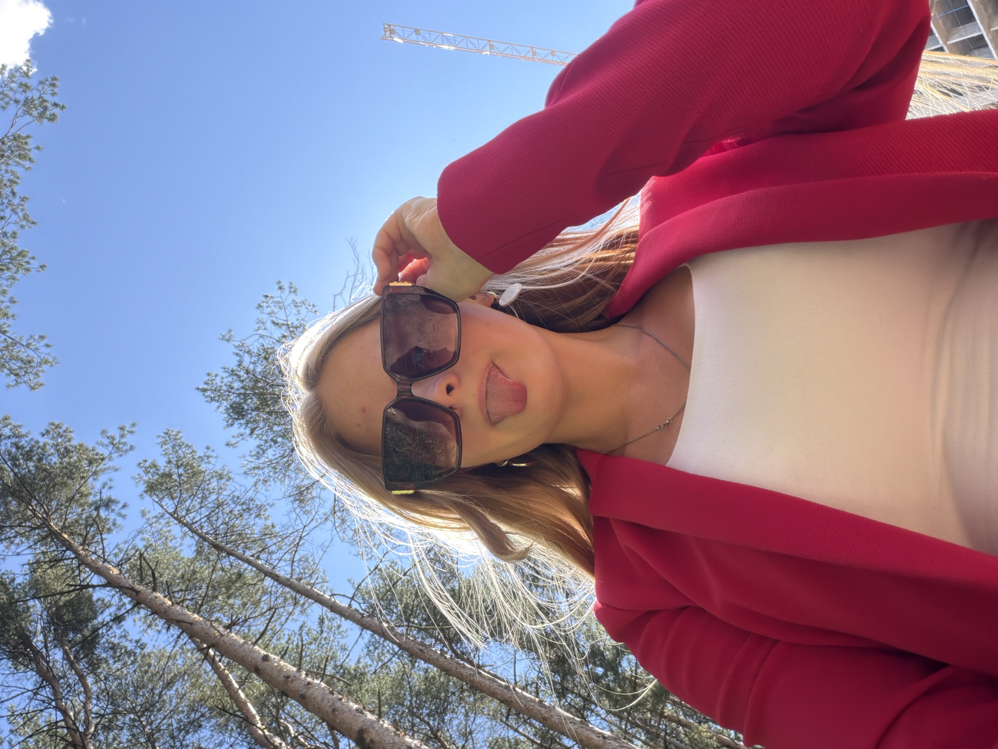A young woman with blonde hair, wearing oversized sunglasses, a white top, and a red blazer, poses for a selfie outdoors. She is sticking her tongue out and adjusting her sunglasses with her right hand. Behind her, tall trees and a clear blue sky are visible, with a construction crane in the distance. The image appears to be taken in Izhevsk, Russia, as indicated by the presence of the construction crane, suggesting an urban or semi-urban environment.