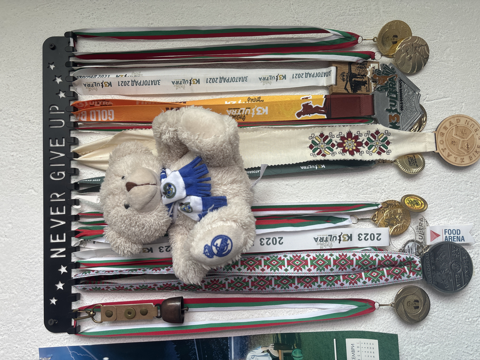 The image captures a collection of athletic achievement medals and ribbons displayed on a black metal hanger against a textured white wall. The hanger features cut-out stars along its left side and the phrase "NEVER GIVE UP" written vertically.
Numerous race ribbons, many showcasing the red, white, and green colors often associated with the Bulgarian flag, are neatly arranged. Some ribbons also exhibit intricate traditional Bulgarian embroidery patterns in similar colors. Attached to these ribbons are various medals, crafted from both metal and wood, indicating participation and completion of different events.
Prominent text visible on the ribbons and medals includes "ZLATOGRAD," "K3ULTRA" (with specific years like 2021, 2022, 2023), "GOLD BORDER RACE," "FOOD ARENA," "FINISHER," "BIKE N RUN," and "MTB." A wooden medal clearly states "ZLATOGRAD BIKE N RUN," while a small golden medal shows the year "2017." Additional Cyrillic text such as "ЗЛАТОГРАД 2021" and "СПОРТЪТ ДА ПОРАСНАТ" (Sport to grow) can also be seen.
A small, light-colored teddy bear, adorned with miniature blue gloves or a scarf featuring a white logo, is nestled among the central ribbons. A similar logo is visible on one of its paws. Below the bear, a small cowbell, attached to a leather strap and ribbon, hangs from the display. At the bottom of the image, a partial calendar is visible, showing "КЕМВРИ" (December) and "СЧПС."
The display signifies a strong commitment to endurance sports, likely trail running or cycling, with multiple events clearly linked to Zlatograd, Bulgaria. The collection spans several years, highlighting consistent participation and achievement.