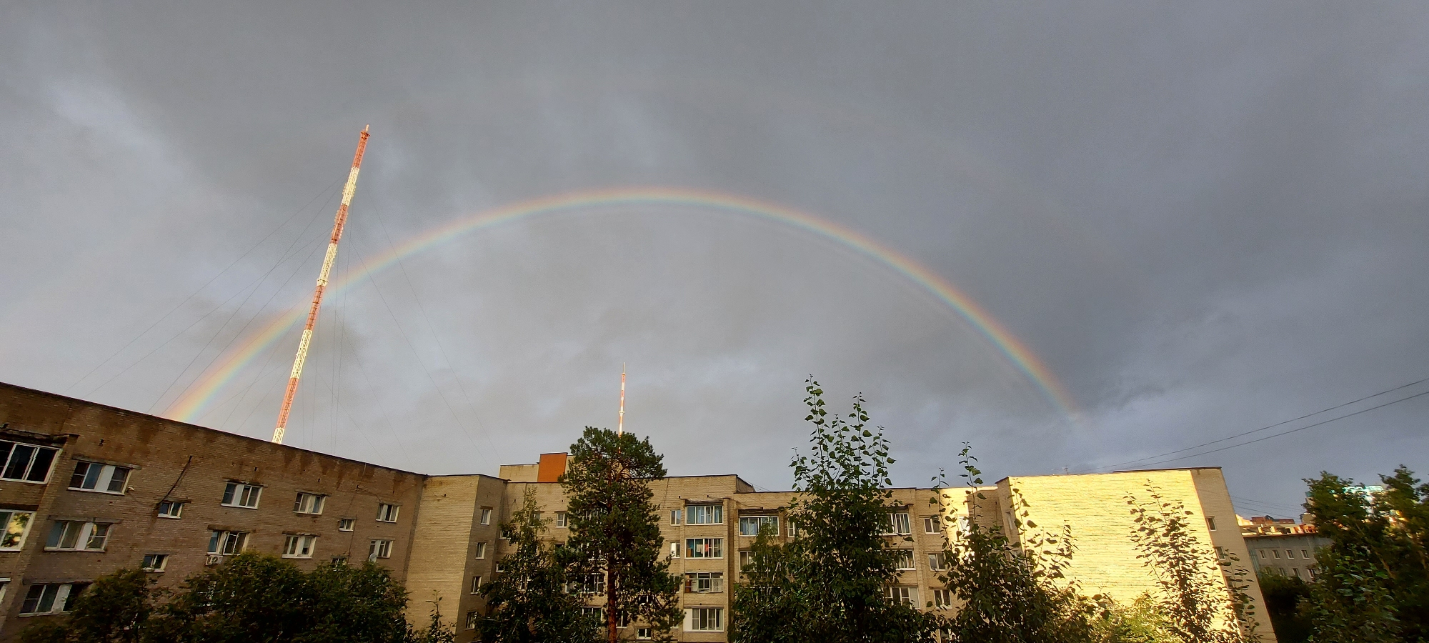 The image depicts a residential building in Chita, Russia, with a rainbow arching across a cloudy sky. The building is constructed of brick and has multiple stories with numerous windows. A tall, striped radio tower stands to the left of the building, partially obscured by the rainbow. Green trees are visible in the foreground and around the building. The sky is overcast with dark clouds, suggesting recent or ongoing rain. The overall scene is indicative of a post-rain urban environment.