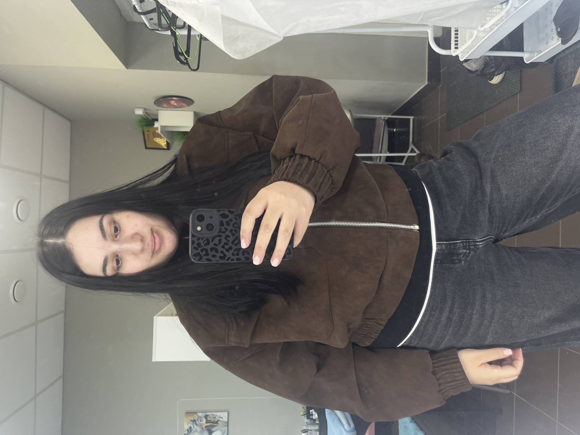 A young woman is captured in a mirror selfie, her reflection filling the majority of the frame. She has long, dark hair, fair skin, and dark eyes, and is looking directly at the camera with a slight smile. Her left hand, as seen in the reflection, holds a smartphone with a black, leopard-print case, positioned to take the picture. Her fingernails are painted with a light pink or nude polish.
She is wearing a brown, suede-like bomber jacket with a silver zipper down the front and ribbed cuffs. Beneath the jacket, a black top or undershirt with a distinct white stripe at the waistband is visible. She pairs this with dark grey or black jeans.
The setting appears to be an indoor commercial space, possibly a salon or hallway. The background, visible in the mirror's reflection, includes light grey walls, a ceiling with white grid lines and circular light fixtures. Various objects are reflected: white shelves or cabinets, a small dark circular decorative item on a wall, and a framed picture with a gold design. Closer to the floor, white shelving units, a black mat, and brown or dark grey tiled flooring are visible. A white, translucent fabric or paper-like material is draped over something in the upper reflected area.
The activity is clearly a mirror selfie. There is no visible text in the image. The time of day and weather are not discernible from this indoor scene. The woman's expression is calm and pleasant.