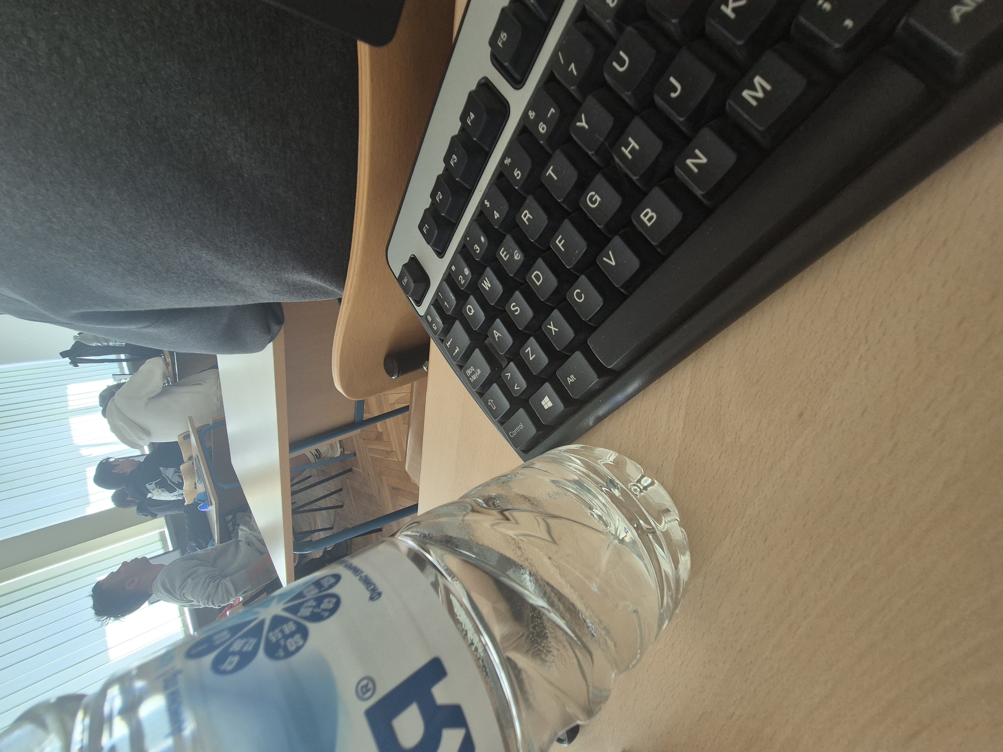 A view from above on a desk setting appears to be taken in Sofia, Bulgaria. A partially visible person wearing a grey garment sits at the desk. A black computer keyboard is positioned on the desk's surface, beside a clear plastic water bottle with a label. In the background, other people are seated at desks, possibly in a classroom or lecture hall. Natural light streams in through a window with blinds. The desks are wooden.