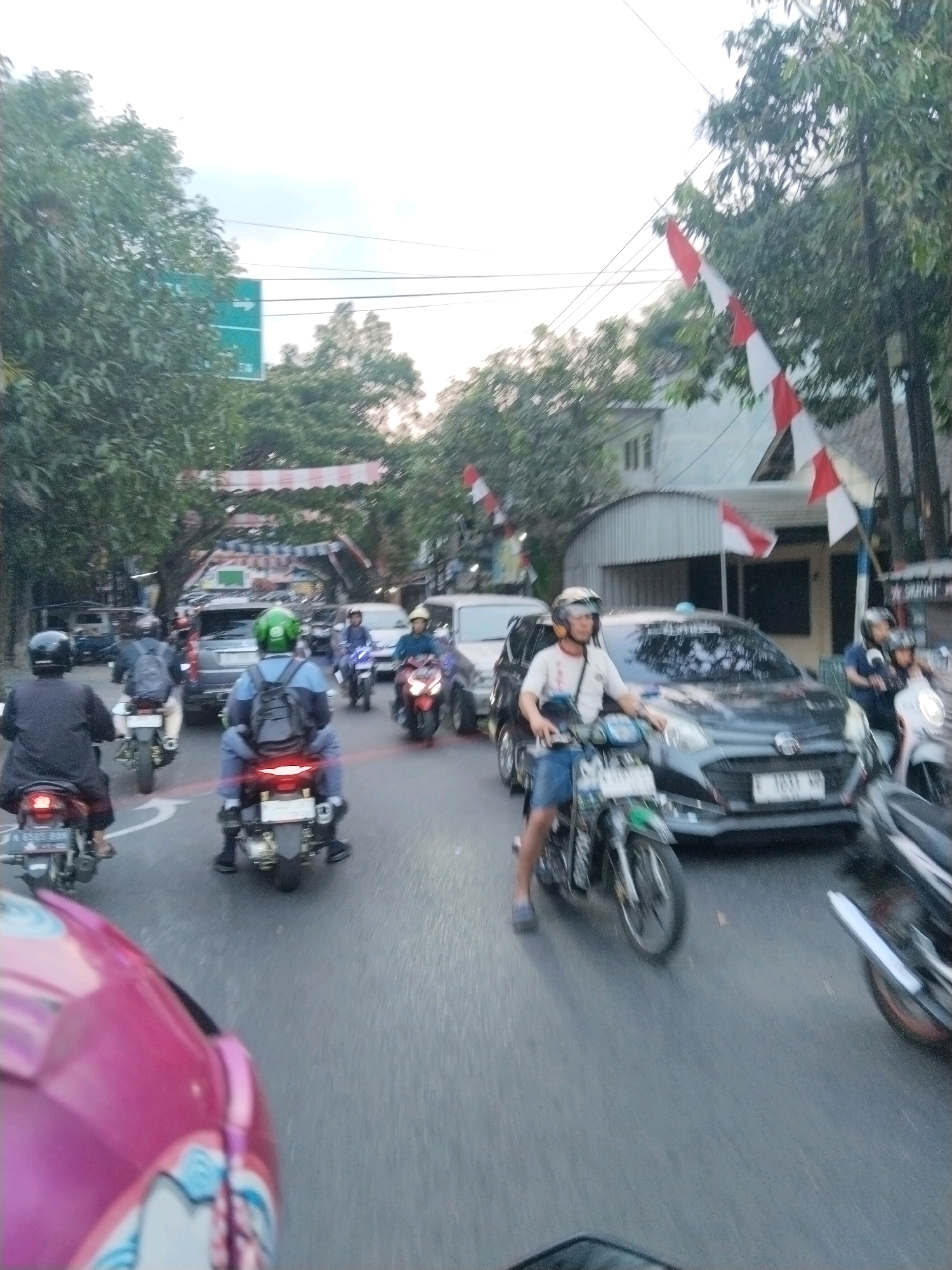This image captures a busy street scene in Malang, Indonesia, likely during daylight. The foreground is dominated by the perspective of a person riding a pink motorcycle, with their helmet partially visible. The road is filled with traffic, primarily motorcycles, with several riders seen wearing helmets. In the middle ground, a dark-colored car is prominent, and other vehicles, including more motorcycles and possibly a van, are also present. The scene is lined with trees, and there are buildings visible in the background, some with awnings or covered structures. Decorative red and white flags, reminiscent of Indonesian national colors, are strung across the street in several places, suggesting a festive or commemorative occasion. A green traffic sign with a white arrow pointing right is visible to the left, indicating directions. The overall atmosphere suggests urban traffic with local cultural elements.