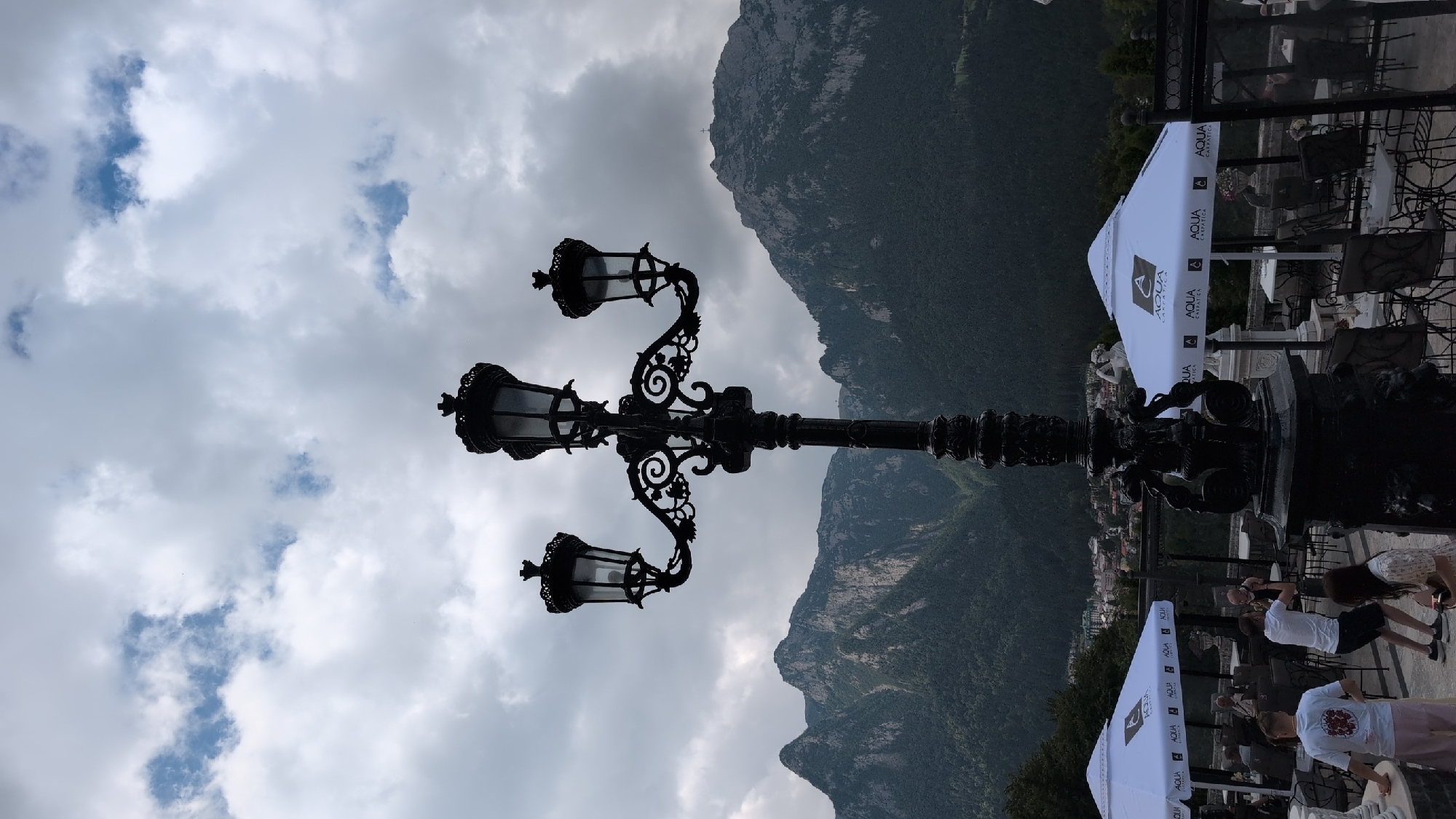 The image shows a view from a terrace in Bușteni, Romania, with a prominent black ornate lamppost in the foreground. In the background, a range of mountains covered in dense green forest rises, with rugged, rocky peaks visible in the upper portions. The sky is overcast with dramatic, heavy clouds.
On the terrace, several white umbrellas with the "AQUA CARPATICA" logo are set up over tables and chairs. Some people are visible, including two men in white t-shirts and shorts standing near the left side of the frame. One man is holding a baby. Another woman is sitting on the right side of the frame. The setting appears to be an outdoor dining area or a viewpoint cafe. The architecture of the terrace and some decorative statues are also partially visible.