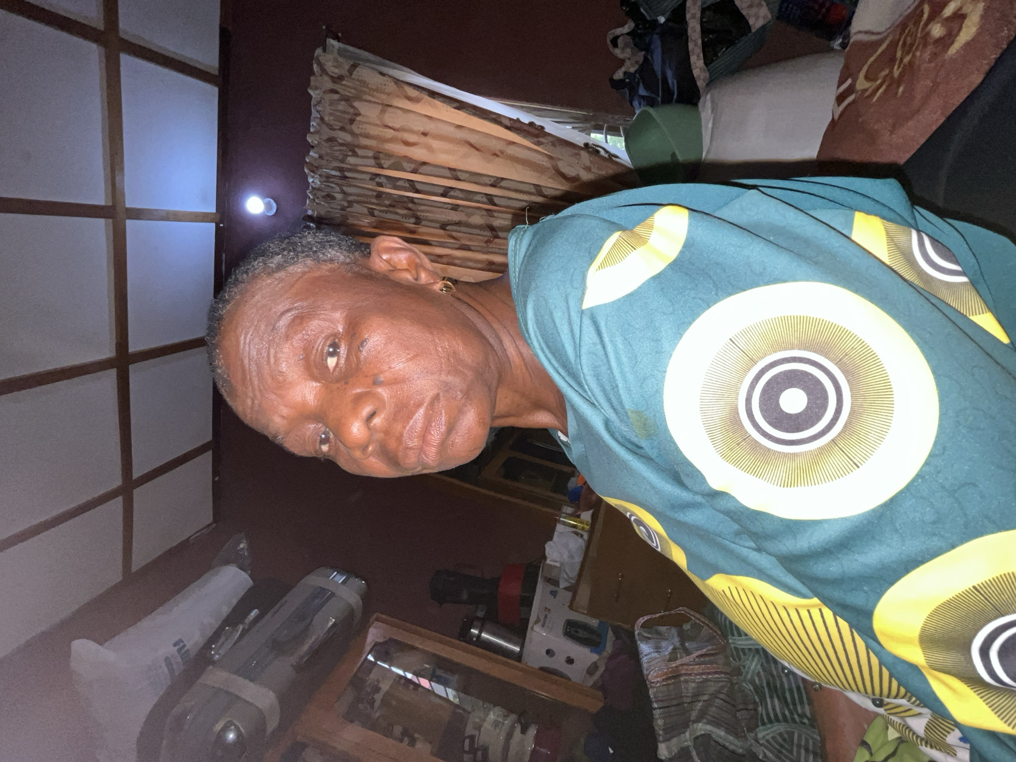 An elderly African woman with short, gray hair is visible from the neck up, wearing a patterned green and yellow shirt. She has dark skin, noticeable wrinkles, and an earring in her left ear. Her gaze is directed towards the viewer. The background includes a combination of domestic elements: a wall with white panels and wooden framing, a light fixture on a darker wall, and patterned curtains. Furniture and other items are partially visible in the lower portion of the image, including what appears to be a mirror, dishes, and household appliances. Based on the context, the location is within a residence in Jos, Nigeria.