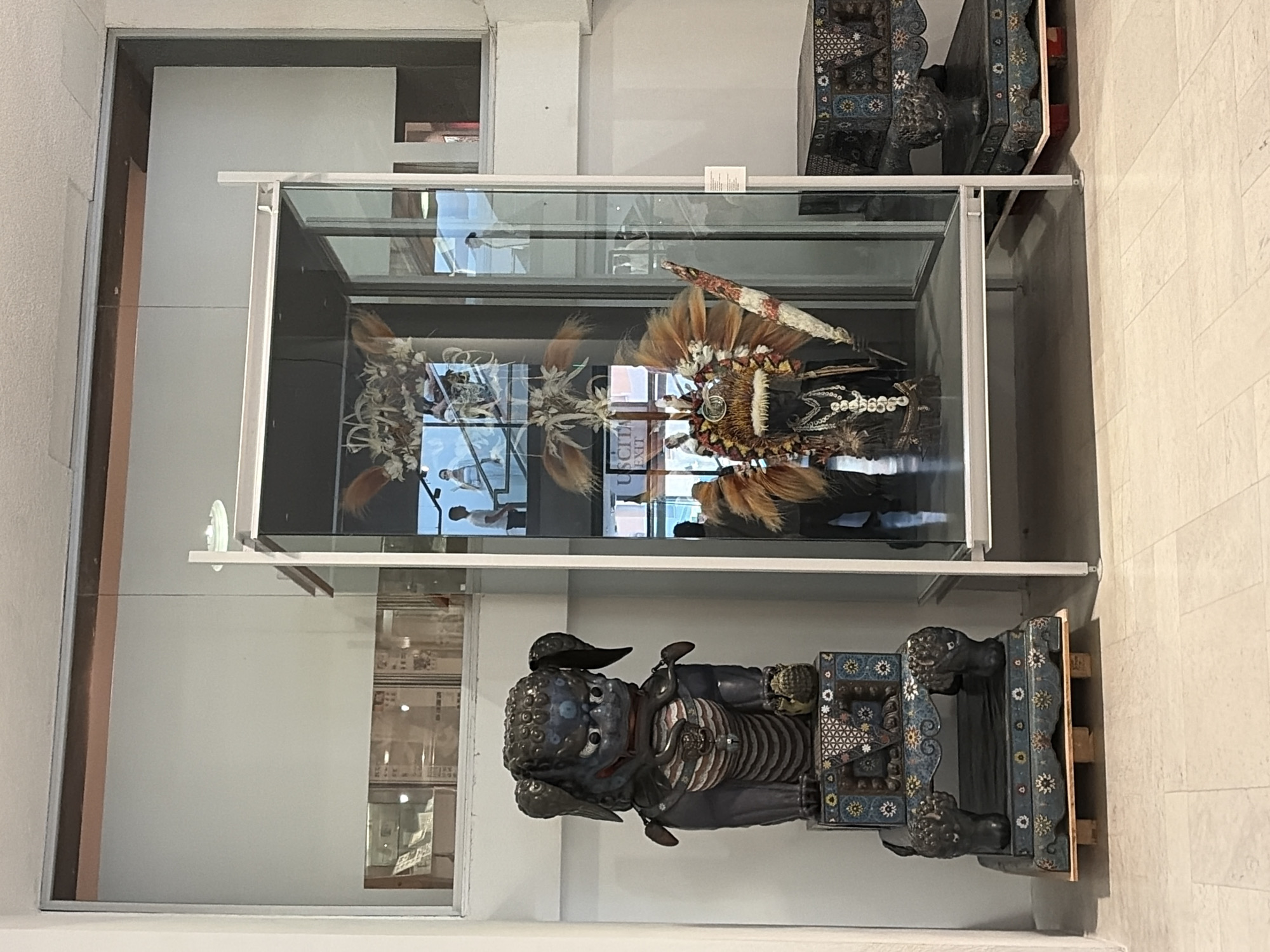 The image shows an interior view of a museum or exhibition space. On the left is a dark blue statue of a mythical creature, possibly a Foo dog, standing on an ornate, multi-tiered pedestal. The creature has a fierce expression, with bulging eyes and fangs. Its body is covered in intricate patterns and what appear to be layered armor. The pedestal is decorated with geometric designs and smaller figurines.
To the right of the statue is a large, glass display case containing two elaborate headdresses or ceremonial costumes. The upper headdress is made of what appears to be bone and feathers, arranged in a fan-like structure. Below it is a second headdress adorned with feathers, beads, and what looks like a wooden staff or spear. Inside the display case, reflections show people viewing the exhibits. A sign in the display case reads "USCITA EXIT."
Further to the right, another smaller, ornate box sits on a pedestal, similar in style to the one supporting the statue. The floor is tiled with light-colored marble. The overall environment suggests a formal exhibition setting with controlled lighting. The image does not provide explicit visual cues to confirm its location within Città del Vaticano, Vatican City.