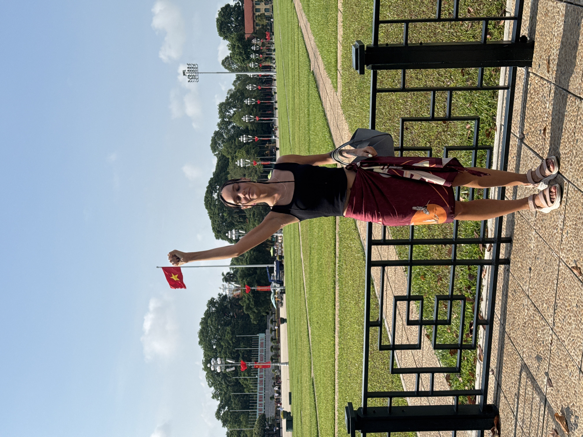 A woman stands on a sidewalk, holding a Vietnamese flag aloft in her right hand. She is wearing a black tank top, a patterned wrap skirt, and sandals. She is standing in front of a black metal fence. Beyond the fence is a large green lawn with a sidewalk running through it. In the background, there are trees, lampposts with Vietnamese flags, and a building. The image appears to be taken at Ba Dinh Square in Hà Nội, Vietnam.