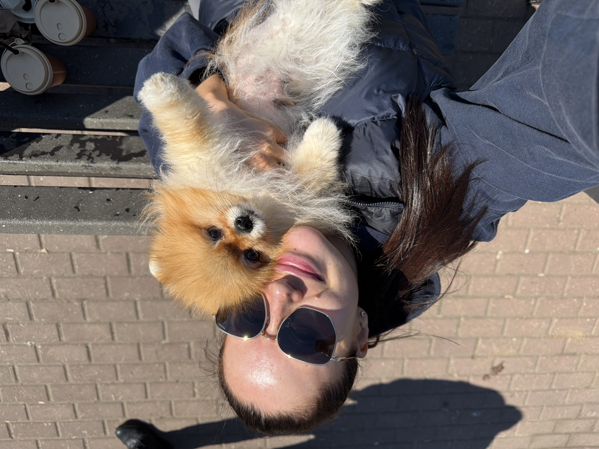 A selfie-style, medium-shot image shows a woman holding a small, orange-colored Pomeranian dog. The woman is wearing sunglasses, and part of her dark hair is visible. She's wearing a dark blue jacket. The dog is facing the camera with its paws held up. Three disposable coffee cups are visible in the upper left corner of the image. The location appears to be outdoors, with a brick-paved surface visible beneath them. Based on an analysis of identifiable buildings and land features, the probable location is Khimki, Russia.