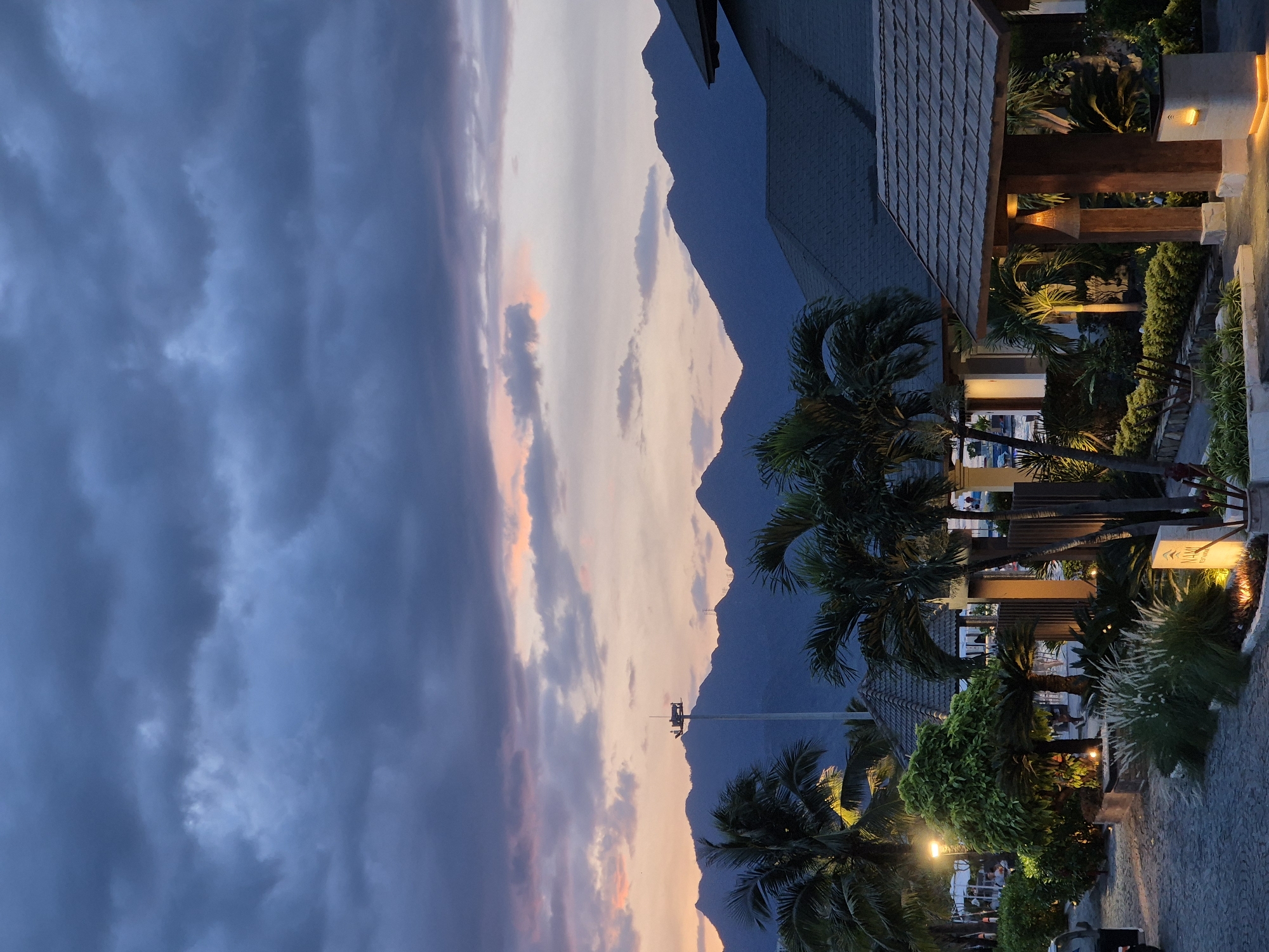 A twilight view captures a resort in Da Nang, Vietnam, featuring a cobbled pathway leading to intricately designed structures. In the foreground, lush greenery and palm trees are illuminated by strategically placed lights, casting a warm glow. Beyond the resort, a range of majestic mountains forms the backdrop, silhouetted against a sky painted with the soft hues of sunset and dramatic, dark clouds. The overall atmosphere is serene and tranquil, with a sense of approaching evening.