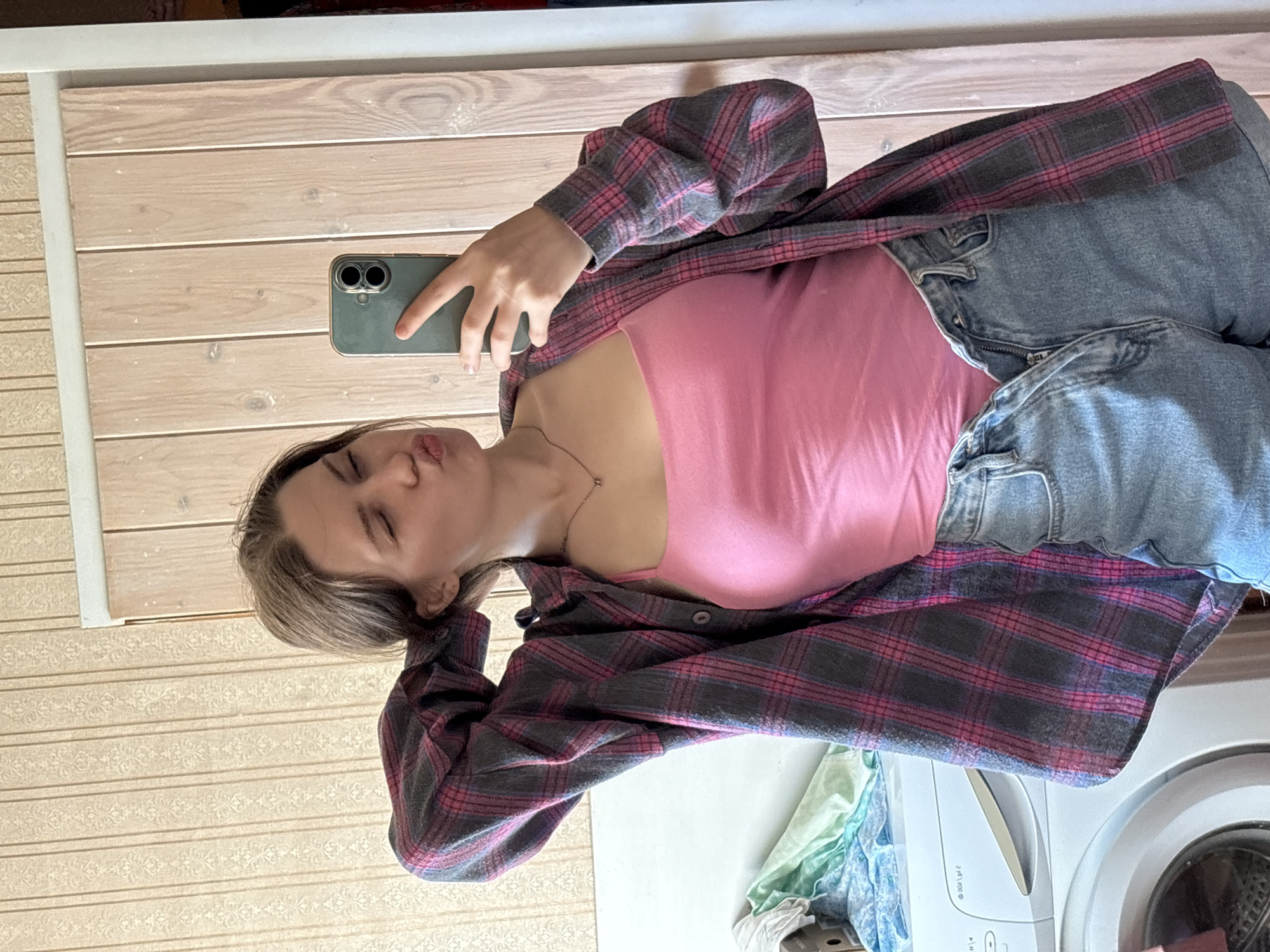 A young woman with brown hair is taking a mirror selfie in a room that appears to be located in Дресвяново, Russia. She is wearing a pink tank top, a plaid flannel shirt unbuttoned and open, and denim shorts. Her left arm is raised, resting on the back of her head, and she is puckering her lips, looking at herself in the mirror. Behind her, a washing machine is visible, along with patterned wallpaper and a wooden door.