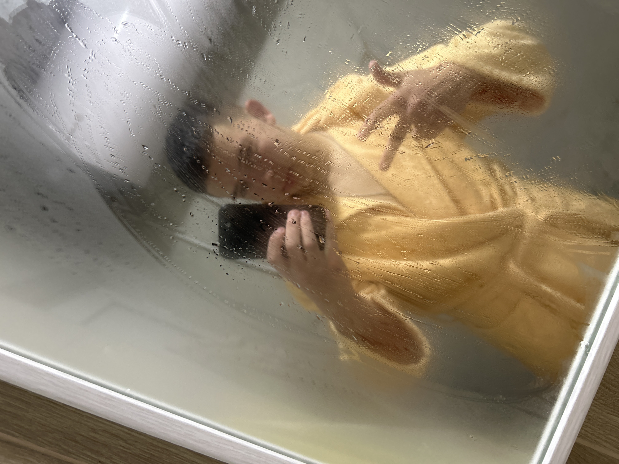 A person is seen through a translucent, steamed-up glass partition, heavily covered with water droplets, creating a distorted and blurred view. The individual appears to be lying on their back inside a shower or bathtub. They are wearing a bright yellow robe or towel.
The person's face is partially visible, turned slightly to the side, and softened by the condensation. One hand is near their face, seemingly holding a dark, rectangular object, possibly a smartphone. The other hand is raised slightly, with fingers spread, resting above their chest area. The pose suggests relaxation or contemplation.
The bottom edge of the image reveals a white trim and a light brown wooden floor, indicating a bathroom setting. The copious water and steam on the glass are a prominent feature, adding an atmospheric quality to the scene. There is no visible text. The image was captured in Zlatograd, Bulgaria.