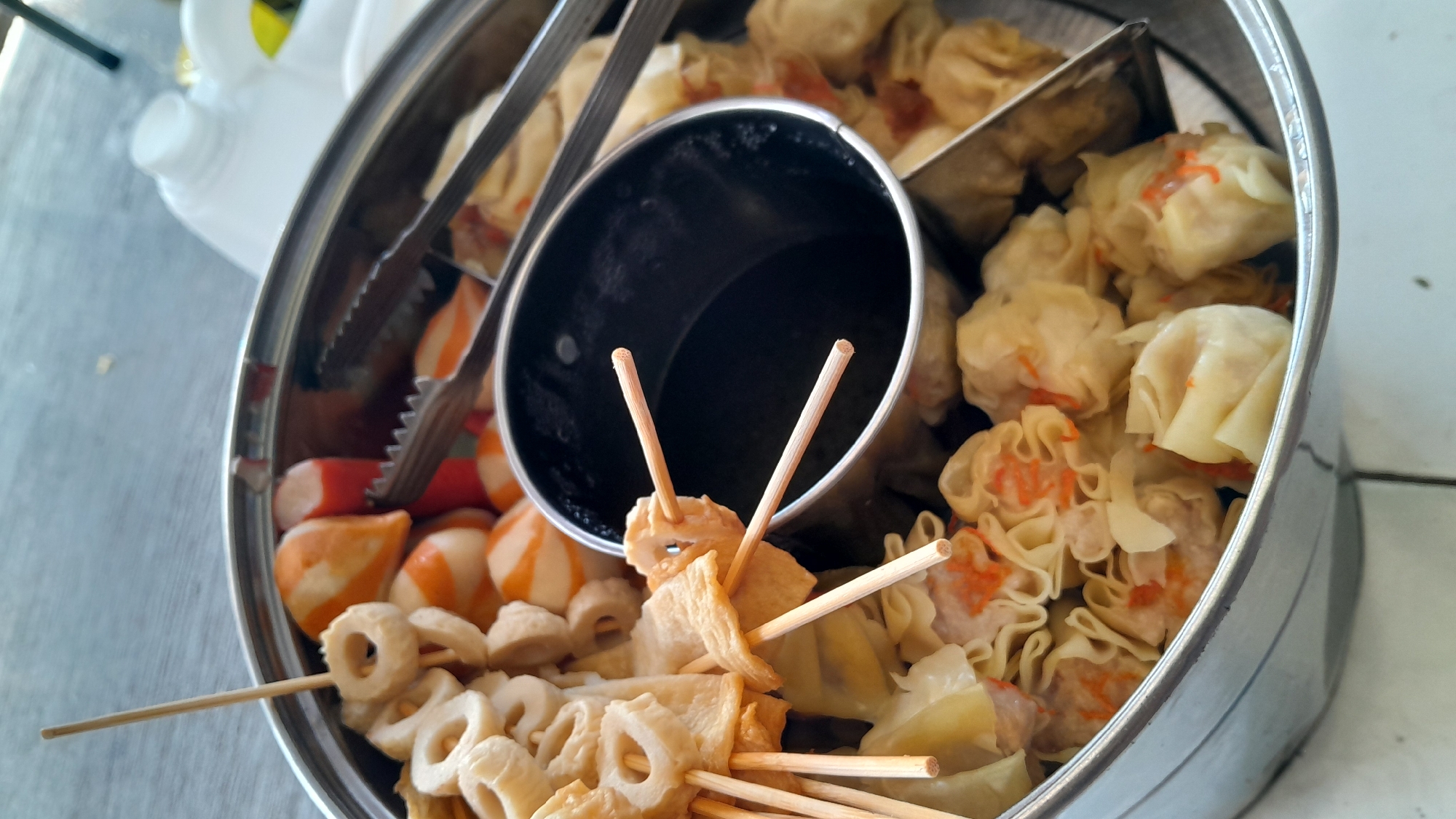 The image shows a stainless steel pot filled with various types of dim sum and fish cakes. The pot is round with a raised rim. Inside, there are several skewers holding pieces of white and orange fish cake, as well as ring-shaped fish cakes. There are also numerous steamed dumplings, likely siomai, visible, some of which have orange flecks of what appears to be shredded carrot. A black metal insert or container is in the center of the pot, possibly for sauce or a broth. A pair of metal tongs rests on the rim of the pot, partially obscuring the dim sum. In the background, a white plastic container and a blurred object are visible. The setting appears to be outdoors, possibly on a table with a textured surface. The overall impression is of a prepared meal, likely ready for consumption. The image does not contain specific visual cues to pinpoint its location to Bandung City, Indonesia.