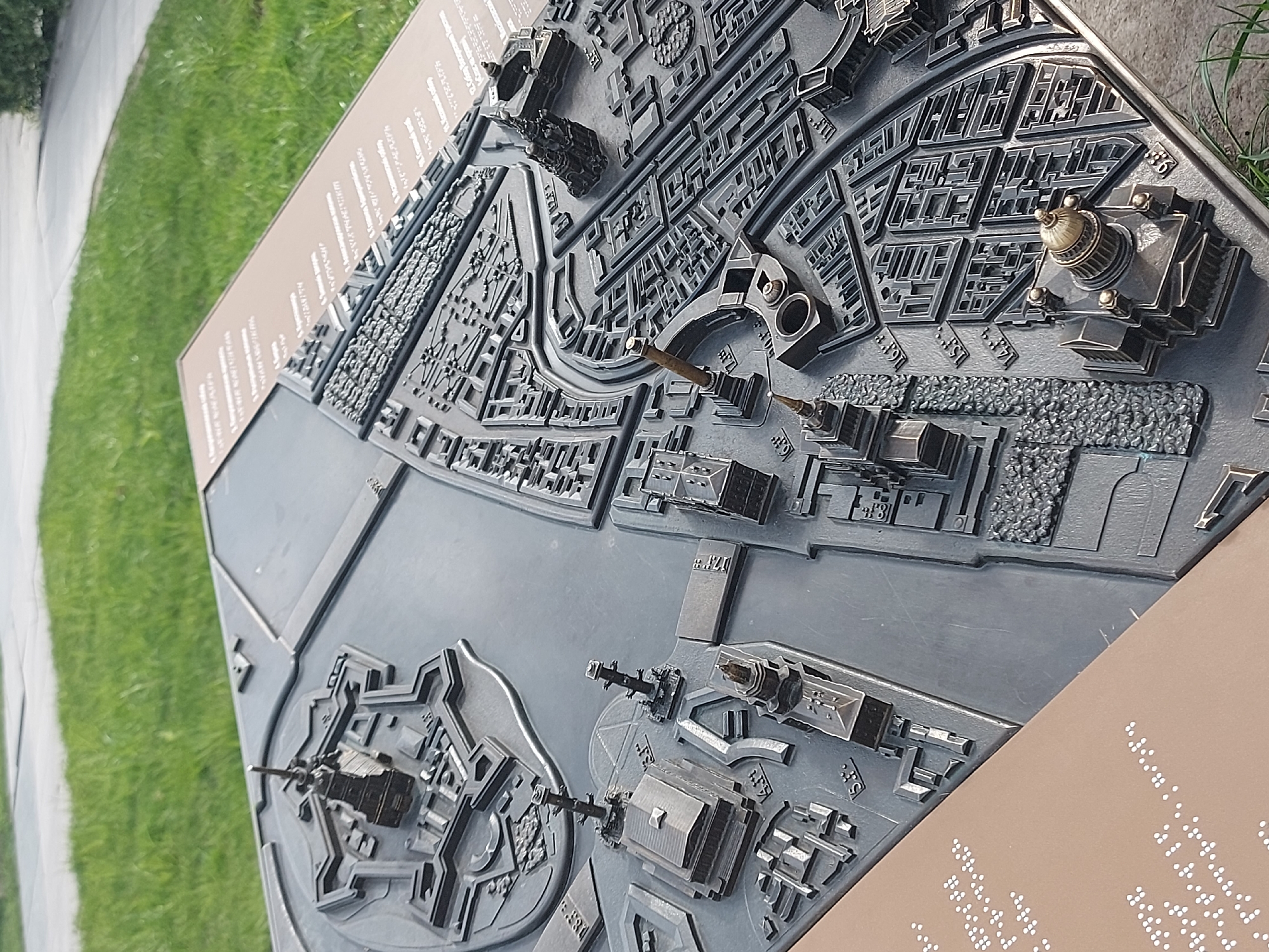 The image shows a raised-relief map of the historical center of St. Petersburg, Russia. The map is oriented at an angle and displays a portion of the Neva River with its prominent historical landmarks. On the left is the Peter and Paul Fortress, a star-shaped bastion, sitting along the Neva River, while to the right is the Winter Palace, a grand, ornate building with a domed structure. The map accurately renders the layout of buildings, streets, and waterways within the historical area. The map is presented on a metal plate mounted on a brown signpost, which includes text in both regular script and Braille. A portion of a grassy area and a concrete walkway are visible on the left side of the signpost.