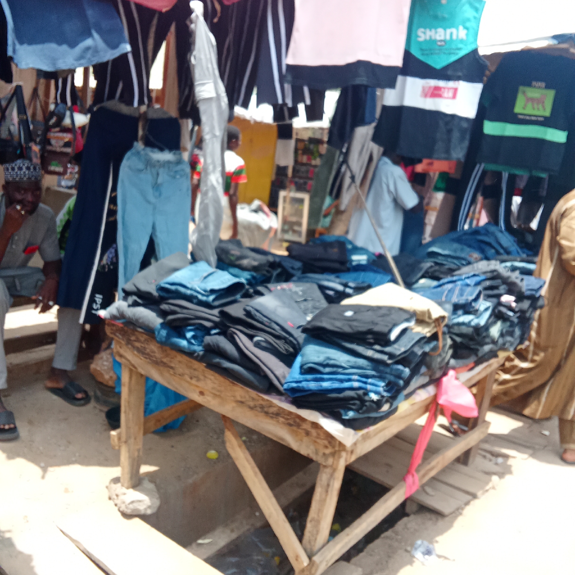 The image depicts a street market scene in an unidentified city. A wooden table is piled with stacks of folded jeans in various shades of blue and black. Clothing, including shirts and pants, hang overhead. Two men are visible: one is seated near the table, wearing a hat, and the other is standing partially out of frame. Additional people are present in the background. The market appears to be outdoors, with a dirt or concrete ground surface.