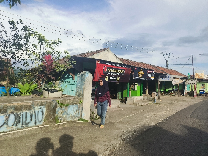 The image shows a street scene in Cikalong, Indonesia (latitude: -6.759, longitude: 107.447). A young woman in a hijab walks along the sidewalk towards the camera. She is wearing a long-sleeved shirt and blue jeans. She is walking past a row of shops with brightly colored awnings. The shops have various signs in Indonesian and English. One shop sign says "DARIM SUPER". Another shop has a sign that says "BENGKEL MOTOR", indicating it is a motorcycle repair shop. There are power lines strung across the street. There is a blue tarp on the ground to the left of the frame. The street is paved and appears to be in good condition. A small, red plant with large leaves grows in a planter box in front of a building. The buildings are all painted in a variety of colors, including blue, green, and yellow. The sky is a light blue with a few clouds. There is a large, bright shadow cast by the building to the left. There is also a shadow of a person walking towards the camera.
