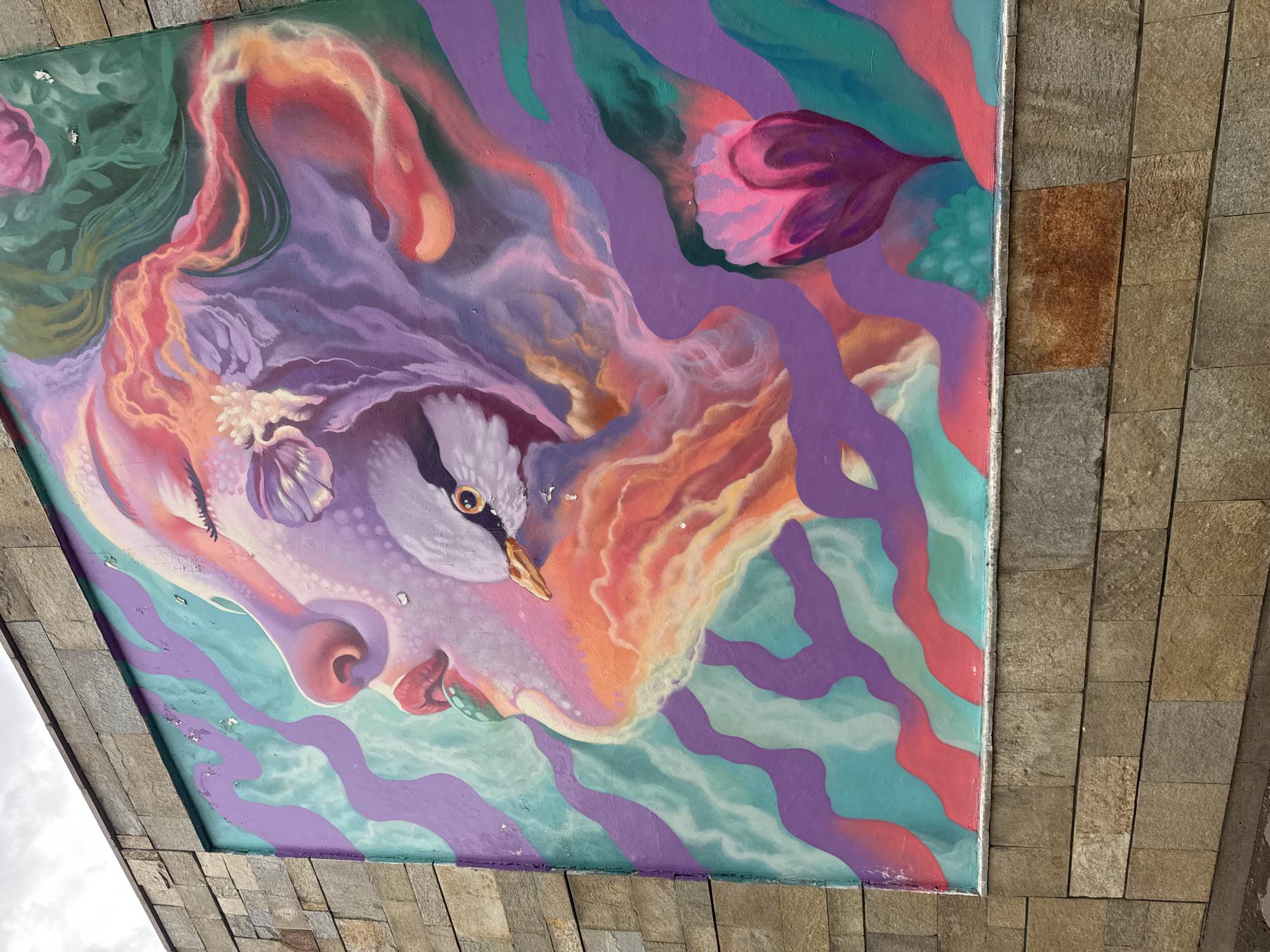 This image shows a mural in Sofia, Bulgaria, located under a bridge or overpass structure. The mural displays an abstract artistic rendering of a human face in profile, with a bird's head as an eye. The colors in the mural are predominantly shades of purple, pink, peach, green, and teal. The surrounding structure is constructed from rectangular, beige stone blocks.