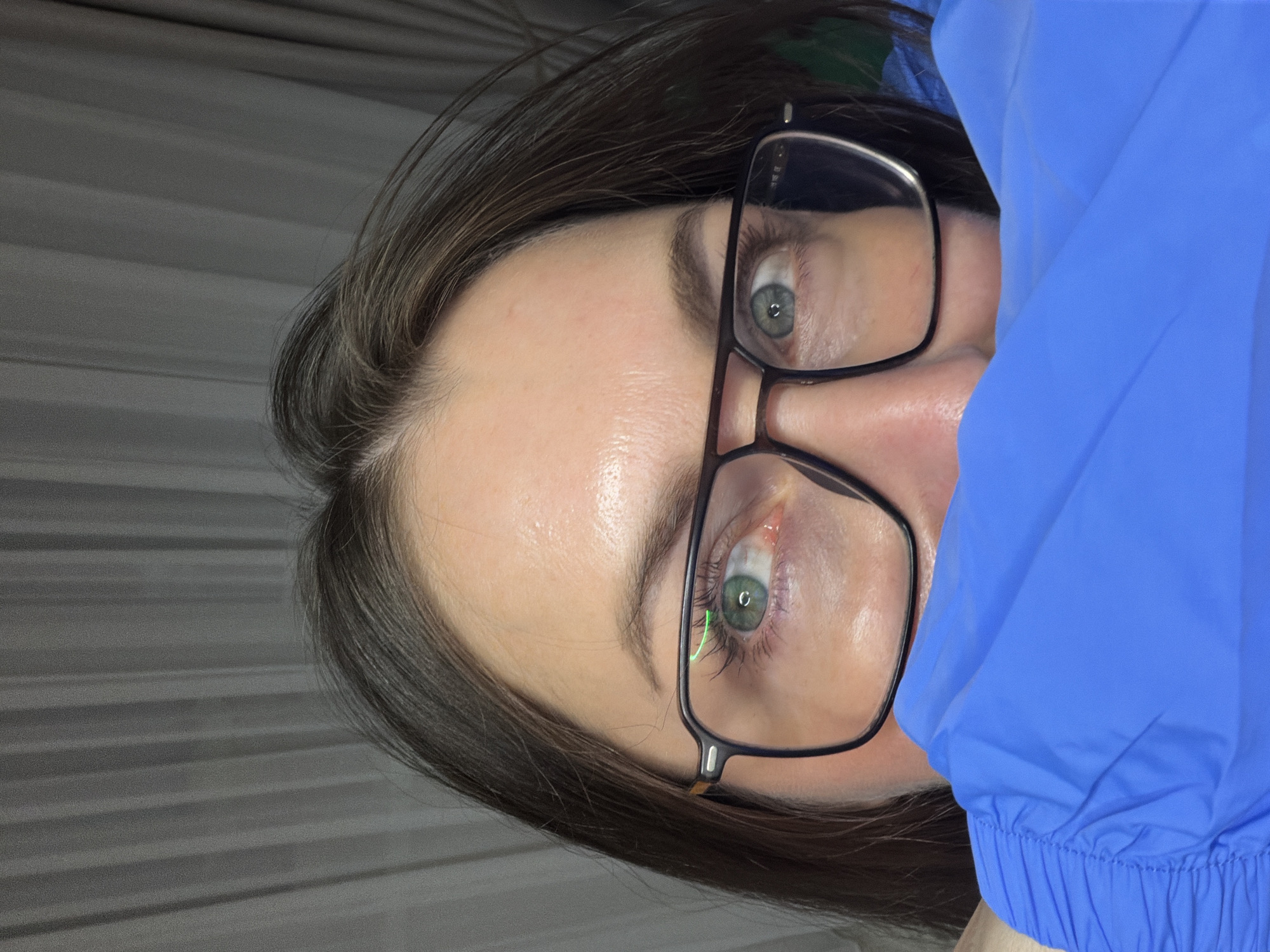 A close-up, slightly angled view of a person wearing glasses and a blue garment. The person has fair skin, dark hair, and light-colored eyes. The glasses have a dark frame. A section of the blue garment covers part of the right side of the face. The background appears to be a gray, textured surface. No location information about Markovo, Russia, is discernible from the image.