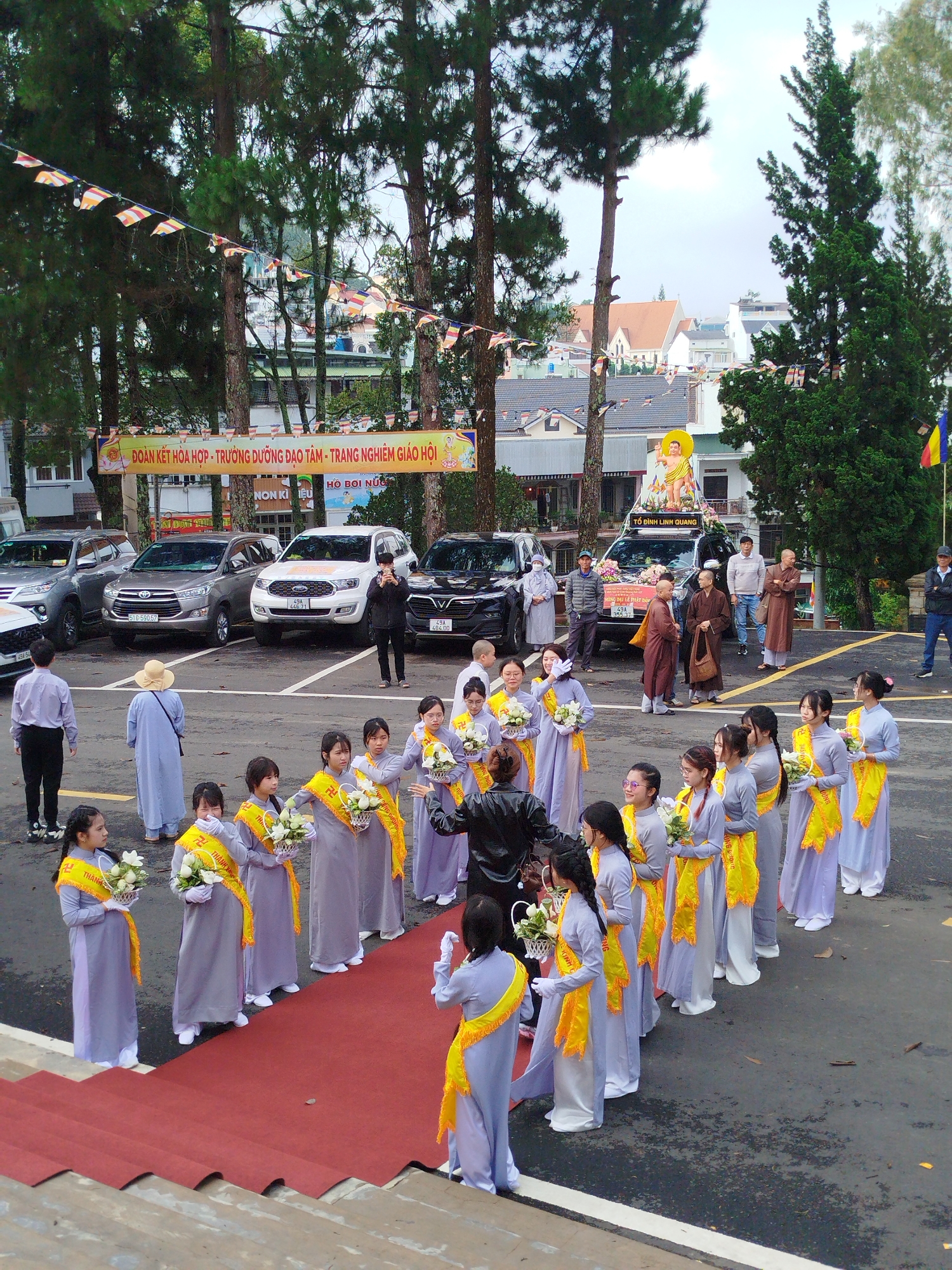 The image shows a ceremony or event taking place in Da Lat, Vietnam. In the foreground, a line of young women dressed in matching light purple outfits with yellow sashes and holding white flowers stand on either side of a red carpet. The sashes appear to have a swastika symbol on them. A person in a dark jacket appears to be directing them.
Behind the women, a group of Buddhist monks in brown robes stand beside a row of parked cars. One of the cars is decorated with a statue of a Buddha figure, indicating a religious significance to the event. A banner with Vietnamese writing hangs above the parked cars.
Several other people are standing in the background, watching the ceremony. The environment includes a paved parking area and trees, with buildings visible in the distance, suggesting an urban or suburban setting within Da Lat. Small pennants are hung between trees.