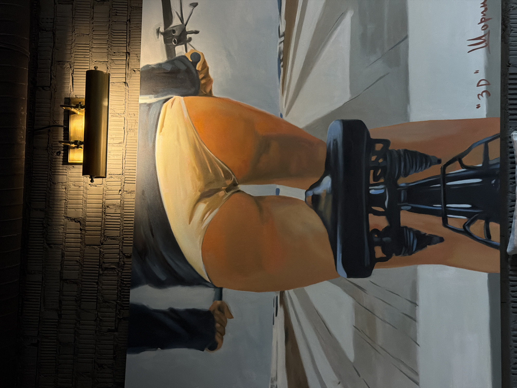 The image shows a painting displayed on a brick wall, illuminated by a brass lamp. The painting depicts a close-up view from behind of a person on a bicycle, wearing light-colored underwear. The cyclist's legs and buttocks are prominently featured, with the rest of the body mostly obscured. The background of the painting suggests an outdoor setting, possibly a road or path stretching into the distance. Red text, reading "'3D' Шорин", is visible in the lower right corner of the painting, indicating the title and artist's name. The overall scene is artistic and suggestive.